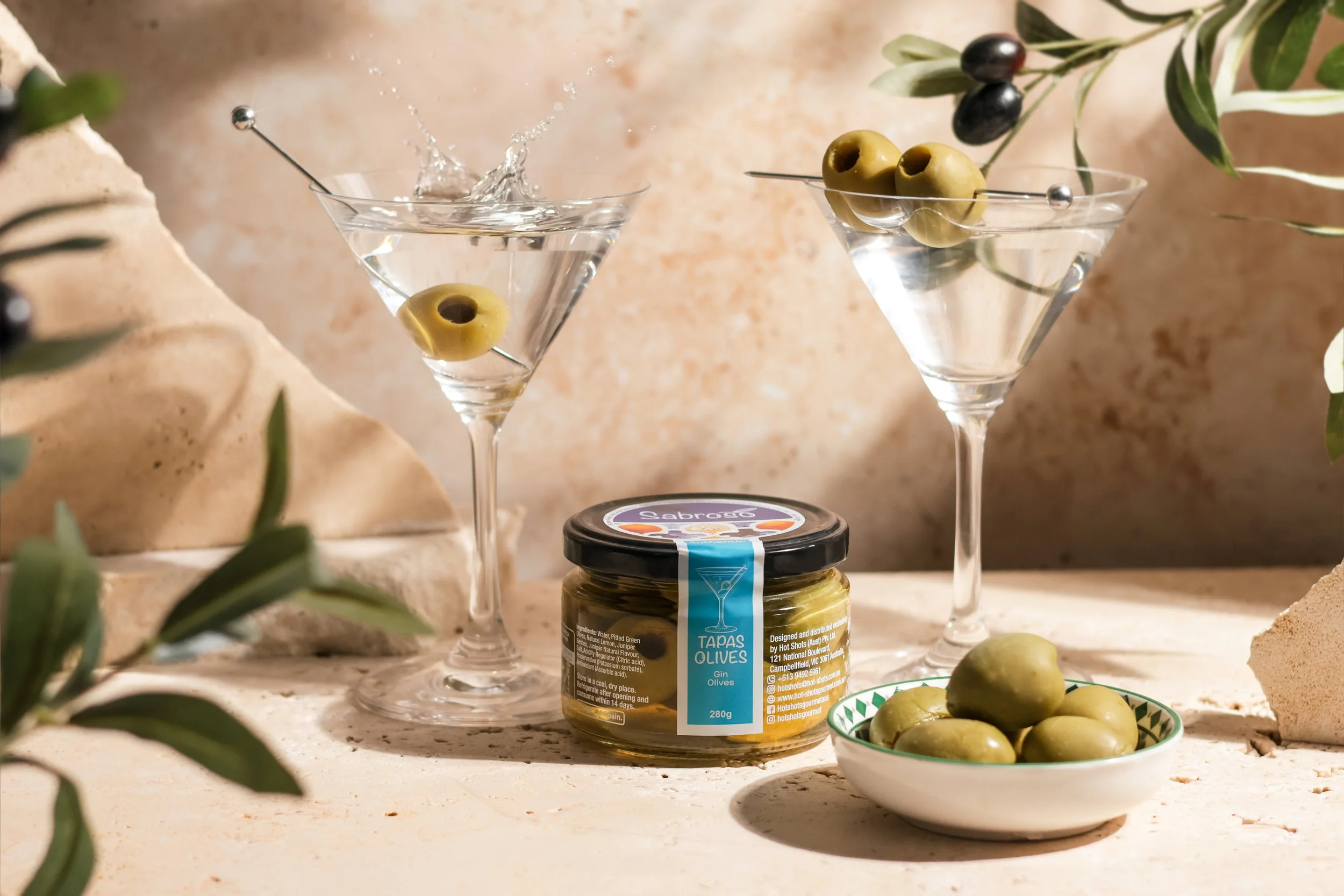 Two martini glasses with green olives, one falling into the glass creating a splash. A jar of green olives with a blue label, a small bowl of more green olives, and some olive branches with black and green olives in the background.