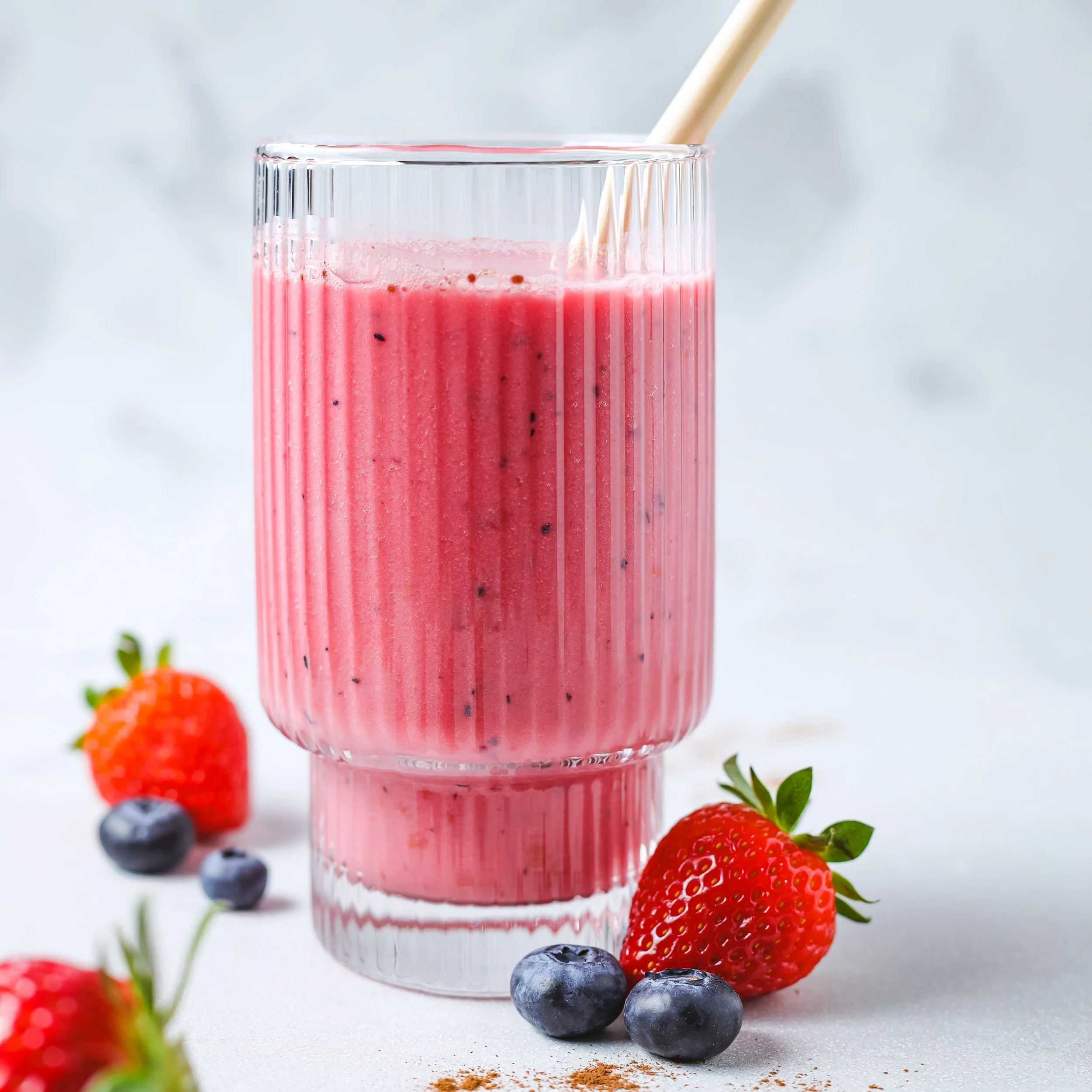 A glass of pink smoothie with a straw, surrounded by fresh strawberries and blueberries on a white surface.