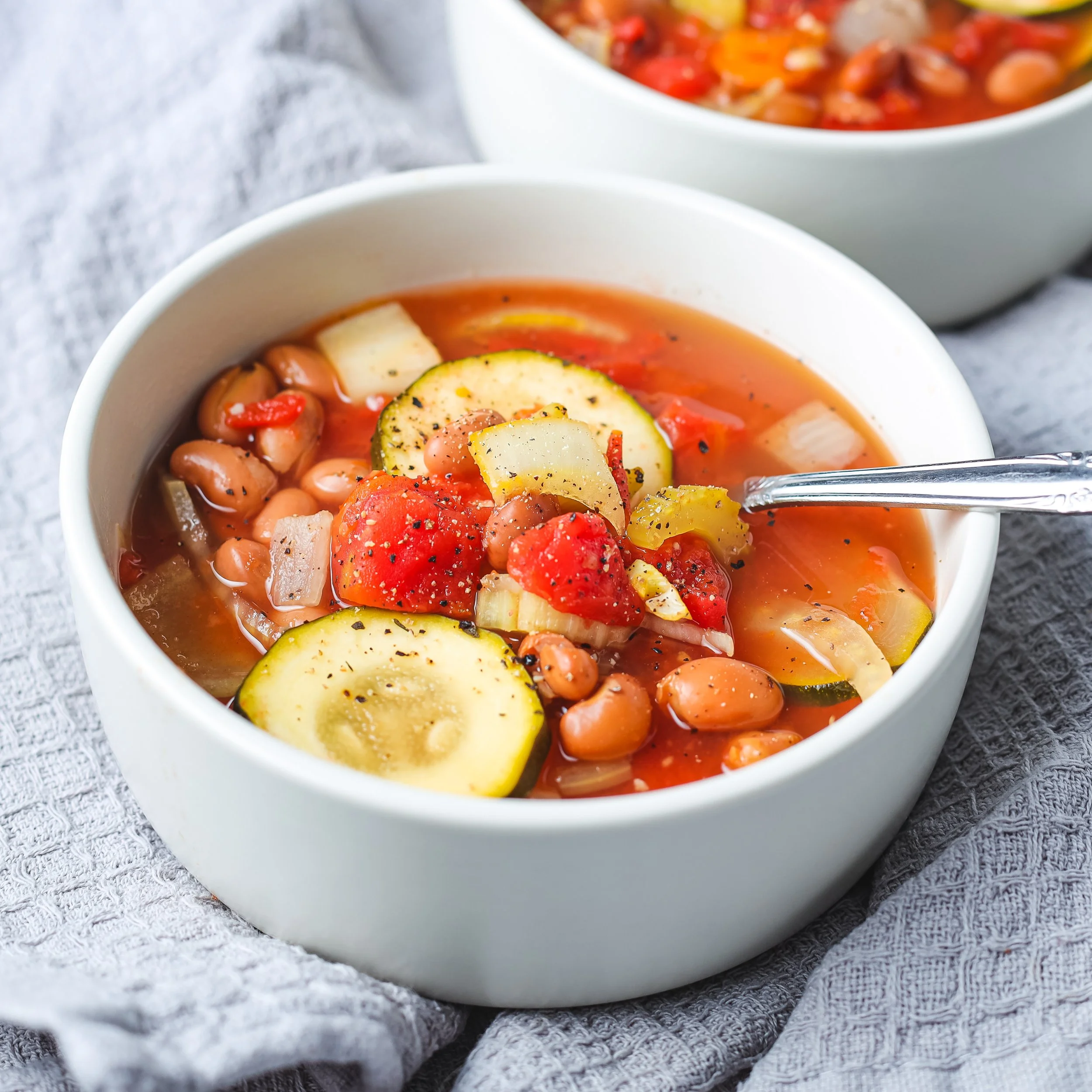 Close-up of a white bowl filled with vegetable soup containing zucchini slices, red tomatoes, beans, and topped with herbs, placed on a grey cloth surface.