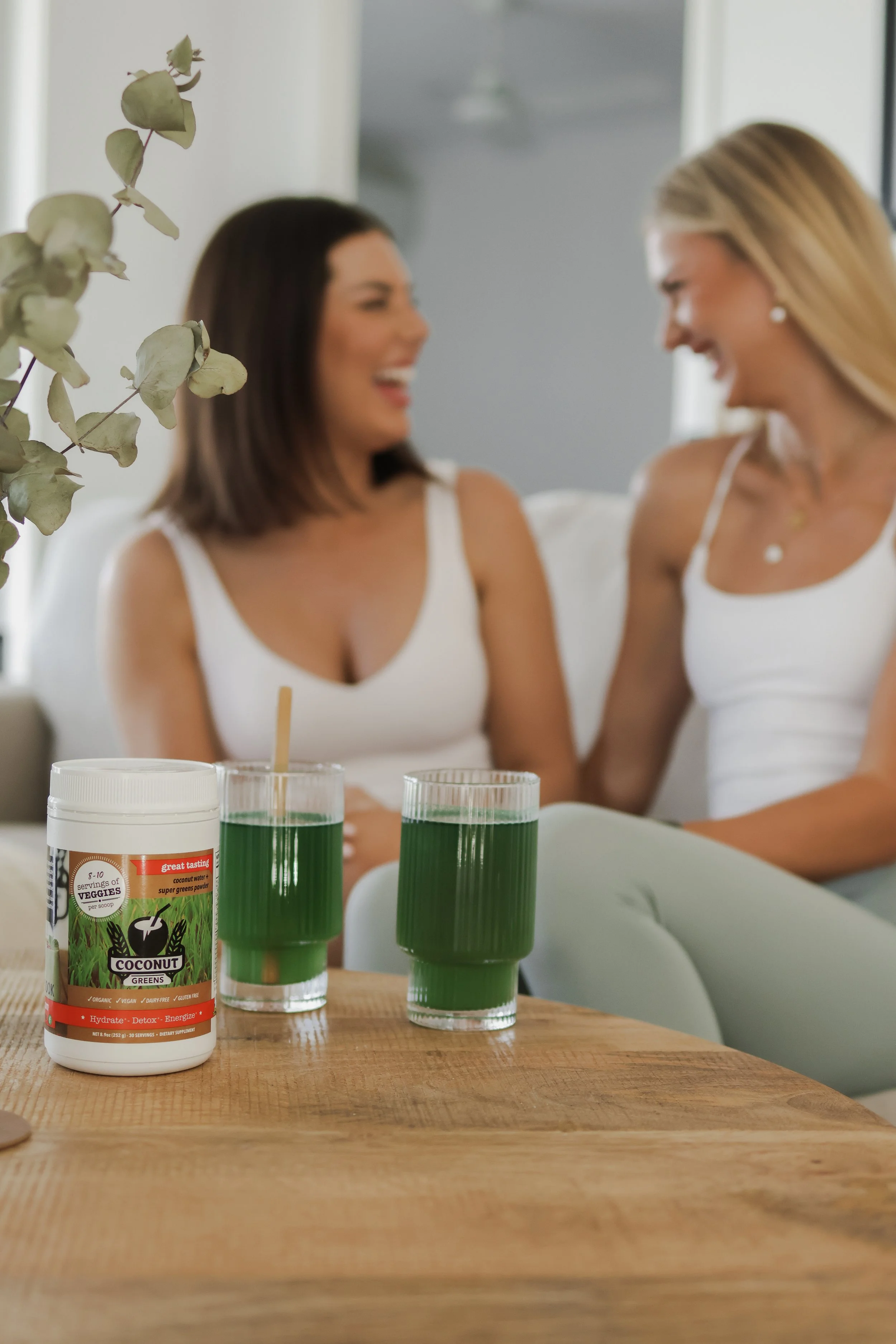 Two women sitting at a table, smiling and laughing, with a front view of a bottle of green superfood powder labeled 'Coconut Greens' and three glasses filled with green liquid in the foreground.