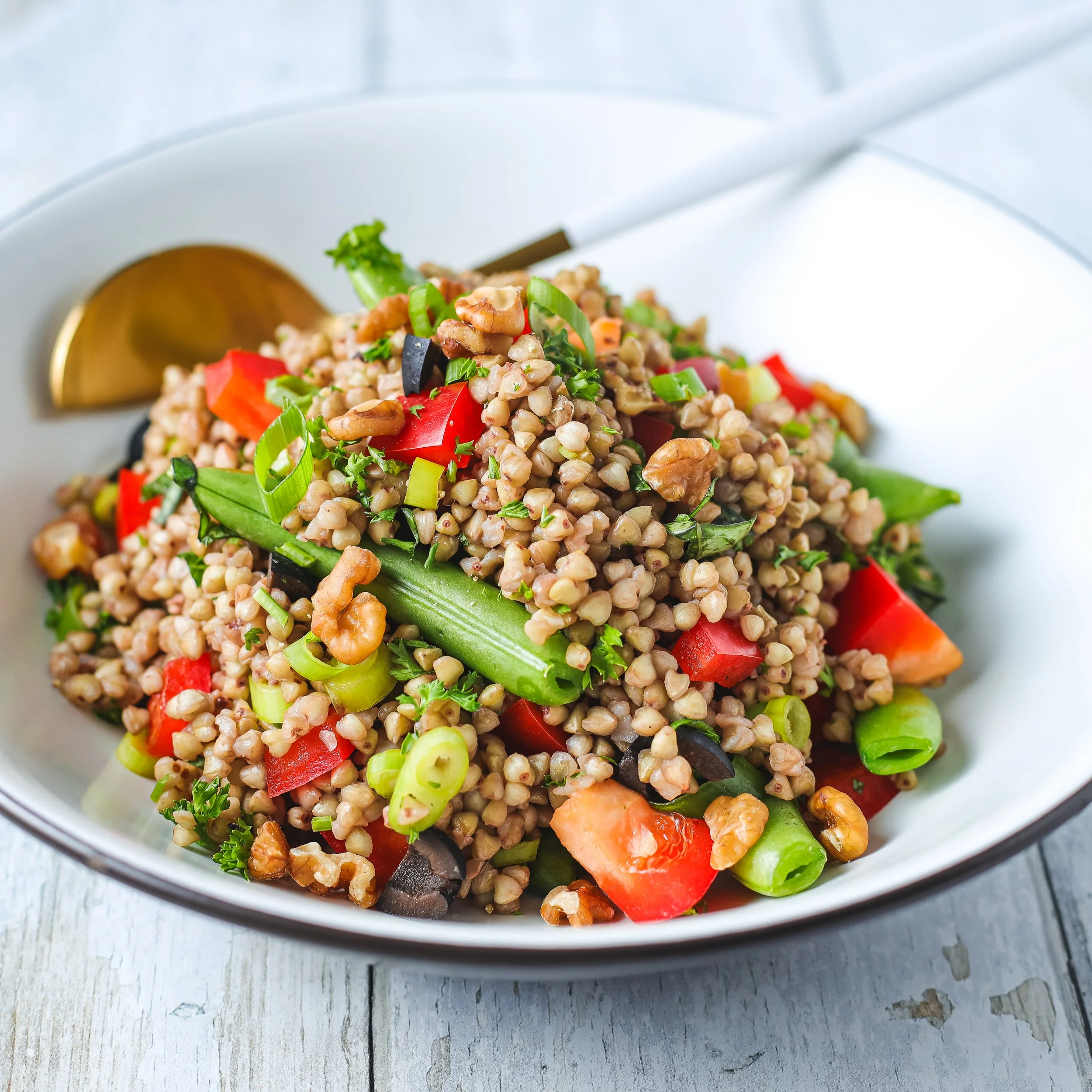 Close-up of a grain salad with wheat berries, red bell peppers, green celery, black olives, walnuts, and parsley in a white bowl on a rustic wooden surface.