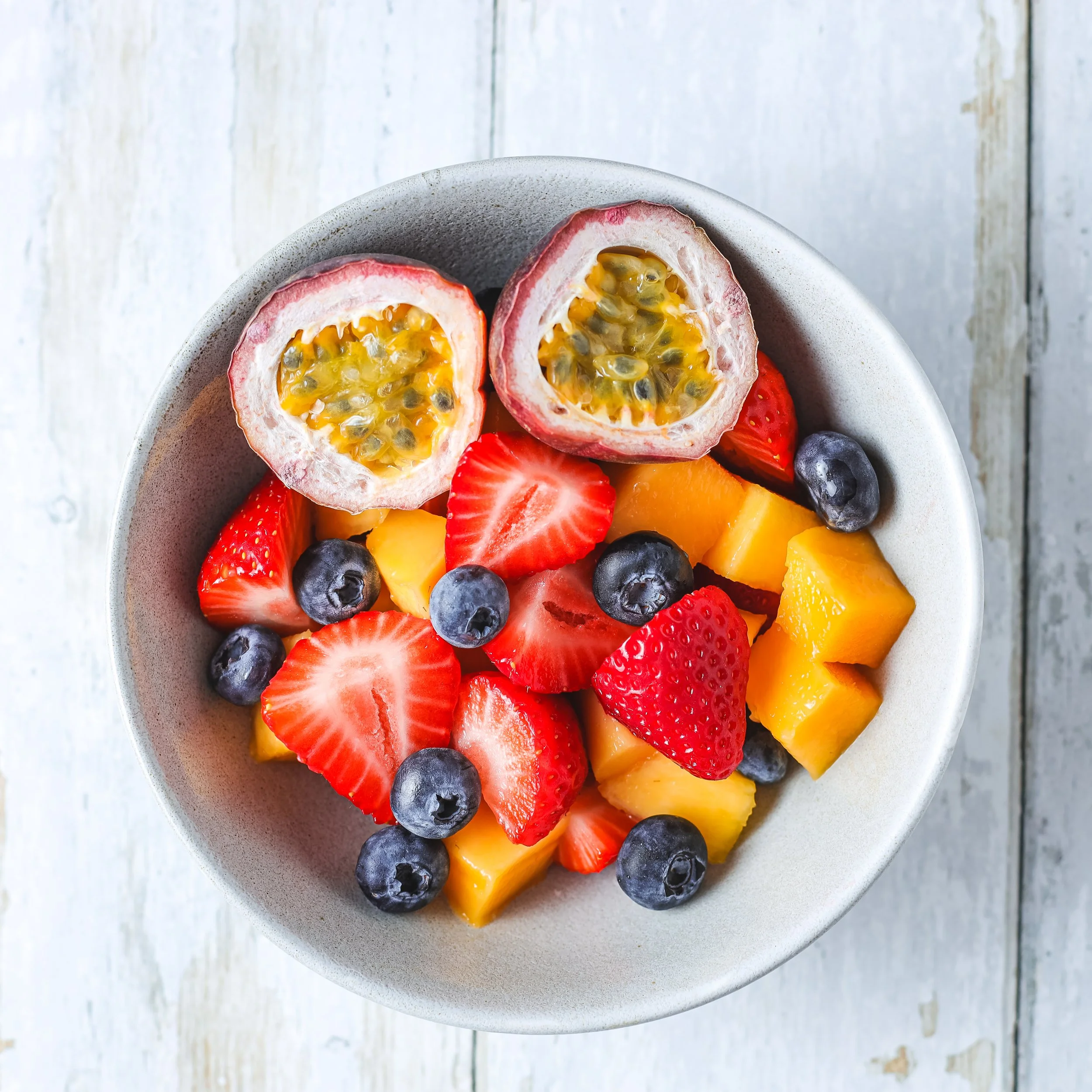 A white bowl filled with various fresh sliced fruits including dragon fruit, strawberries, blueberries, and mango on a white wooden surface.