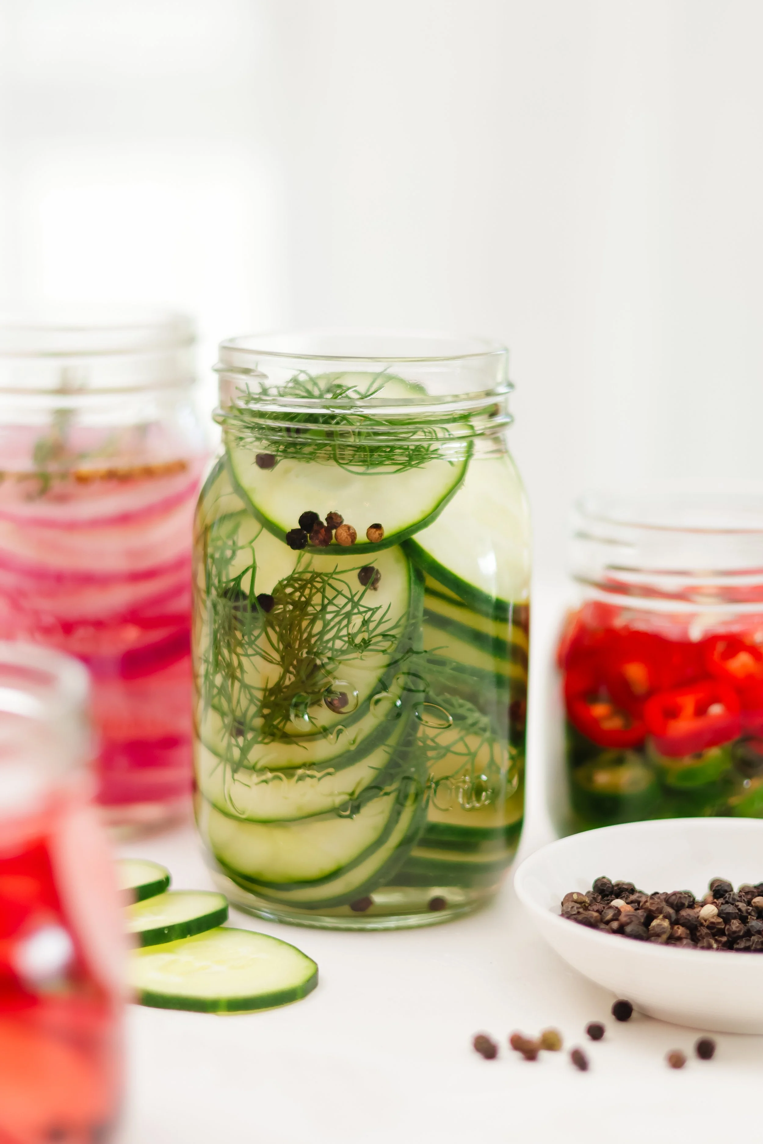 A glass jar filled with cucumber slices, sprigs of dill, and peppercorns on a white surface. Blurred jars with pink and red contents are in the background and foreground.