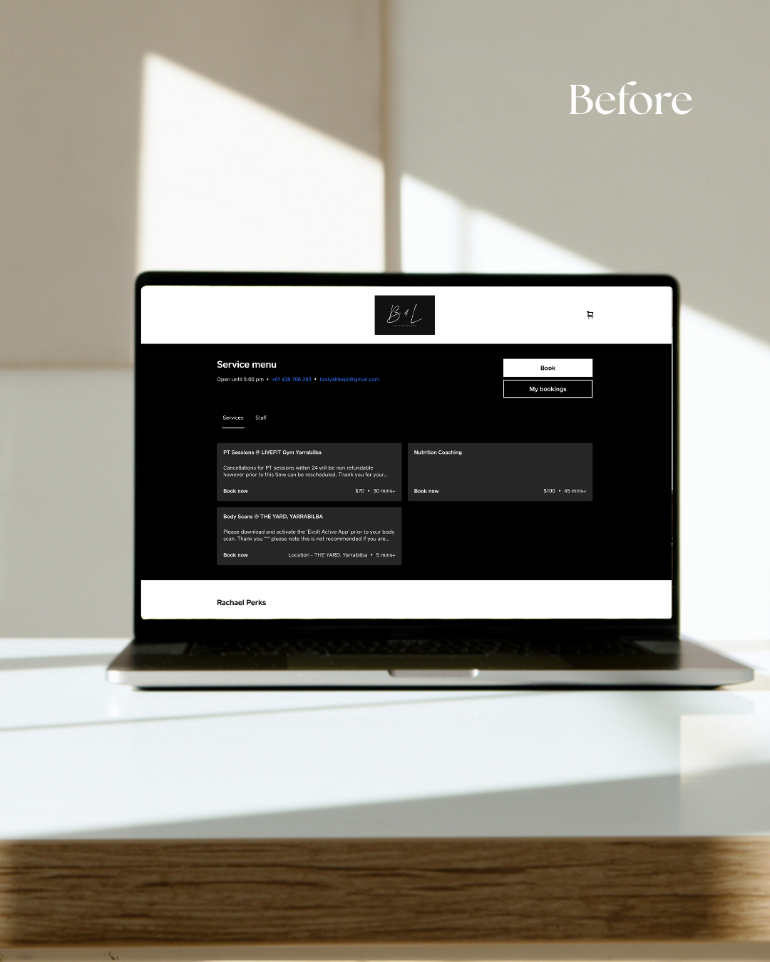 Laptop displaying a website with a black and white service menu, placed on a white desk with a wooden edge, sunlight casting shadows on the wall behind.