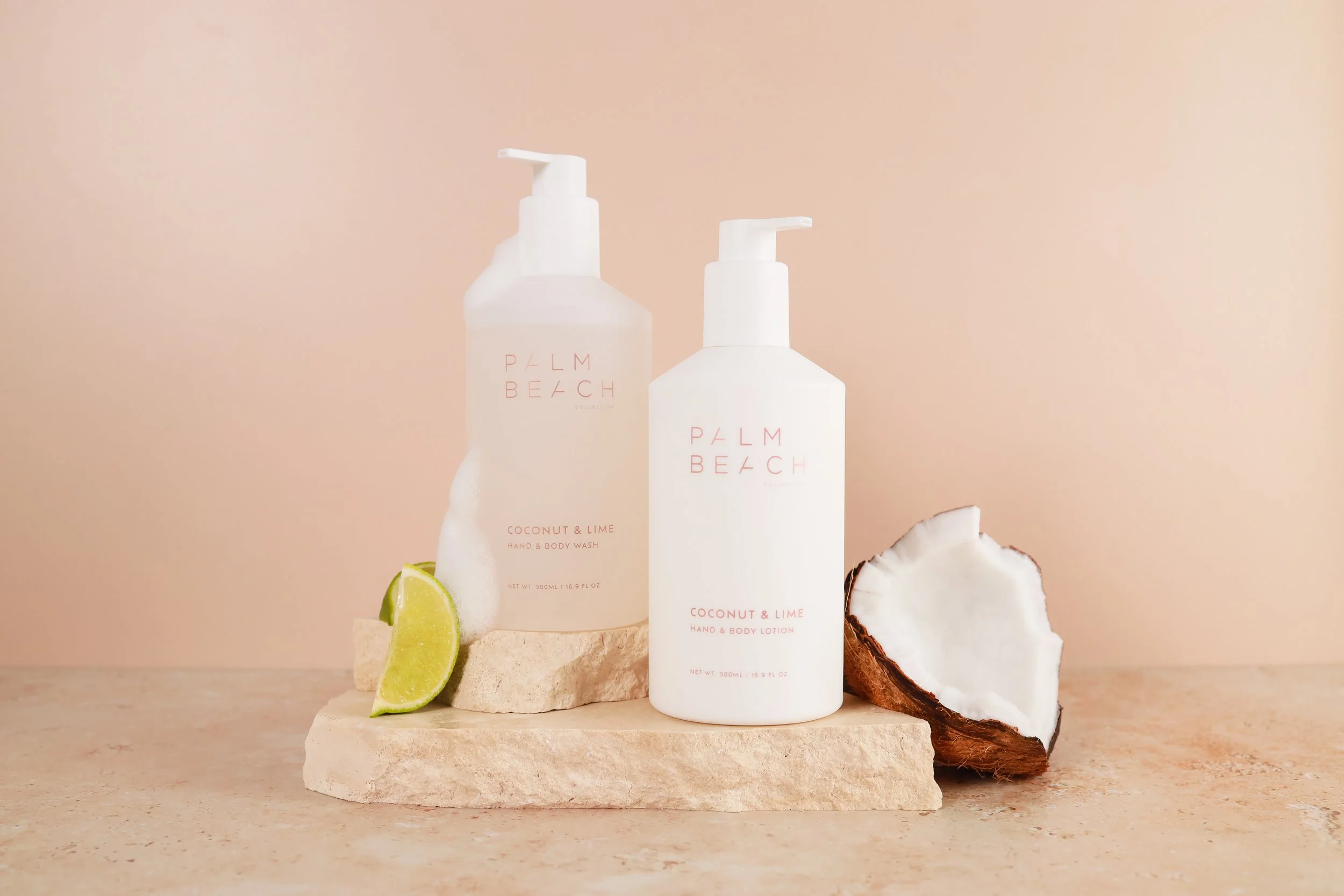 Two white bottles of Palm Beach coconut and lime hand and body wash and lotion on a beige stone slab, with lime wedges and a halved coconut nearby, against a light pink background.