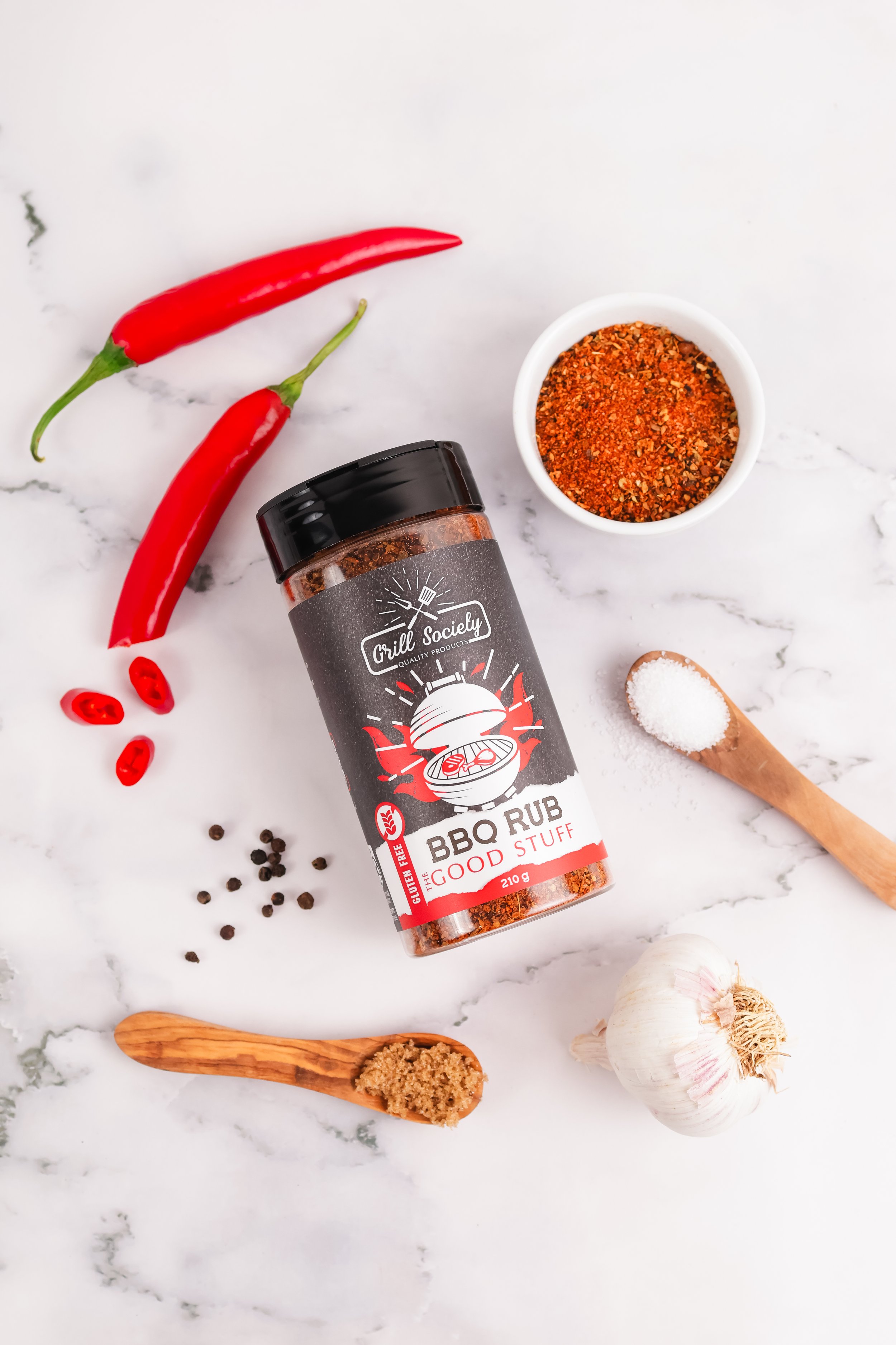 Container of barbecue rub seasoning surrounded by red chili peppers, garlic, black peppercorns, salt, and a wooden spoon on a white marble surface.