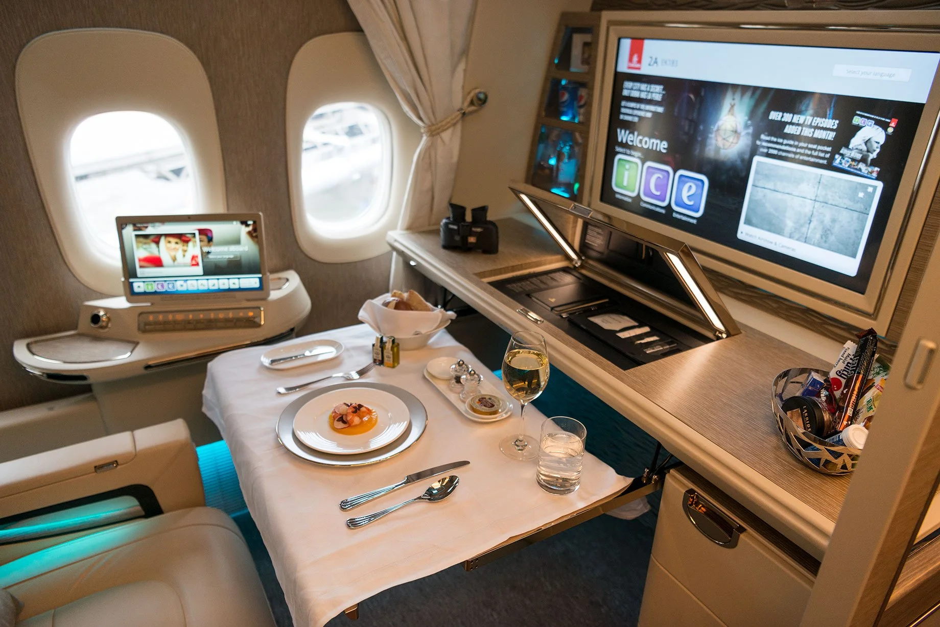Luxury airplane cabin dining setup with a small table, plated meal, glass of wine, TV screen, and private screens near airplane windows.
