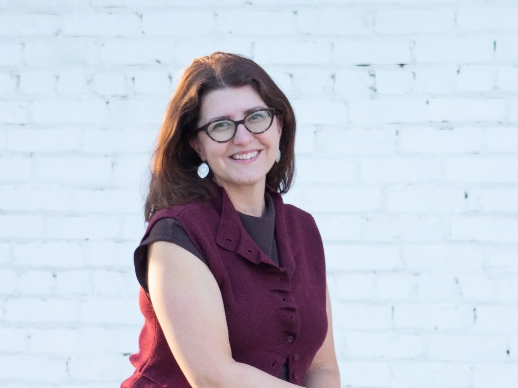 A headshot of Amanda Glatfelter with brown hair, wearing glasses, earrings, and a burgundy vest, smiling and crossing her arms in front of a white brick wall.