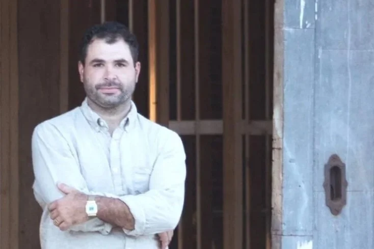 A headshot of Bo Wright with dark hair and a beard, wearing a light-colored button-up shirt, crossing his arms, standing in front of a partly constructed wooden and concrete building.