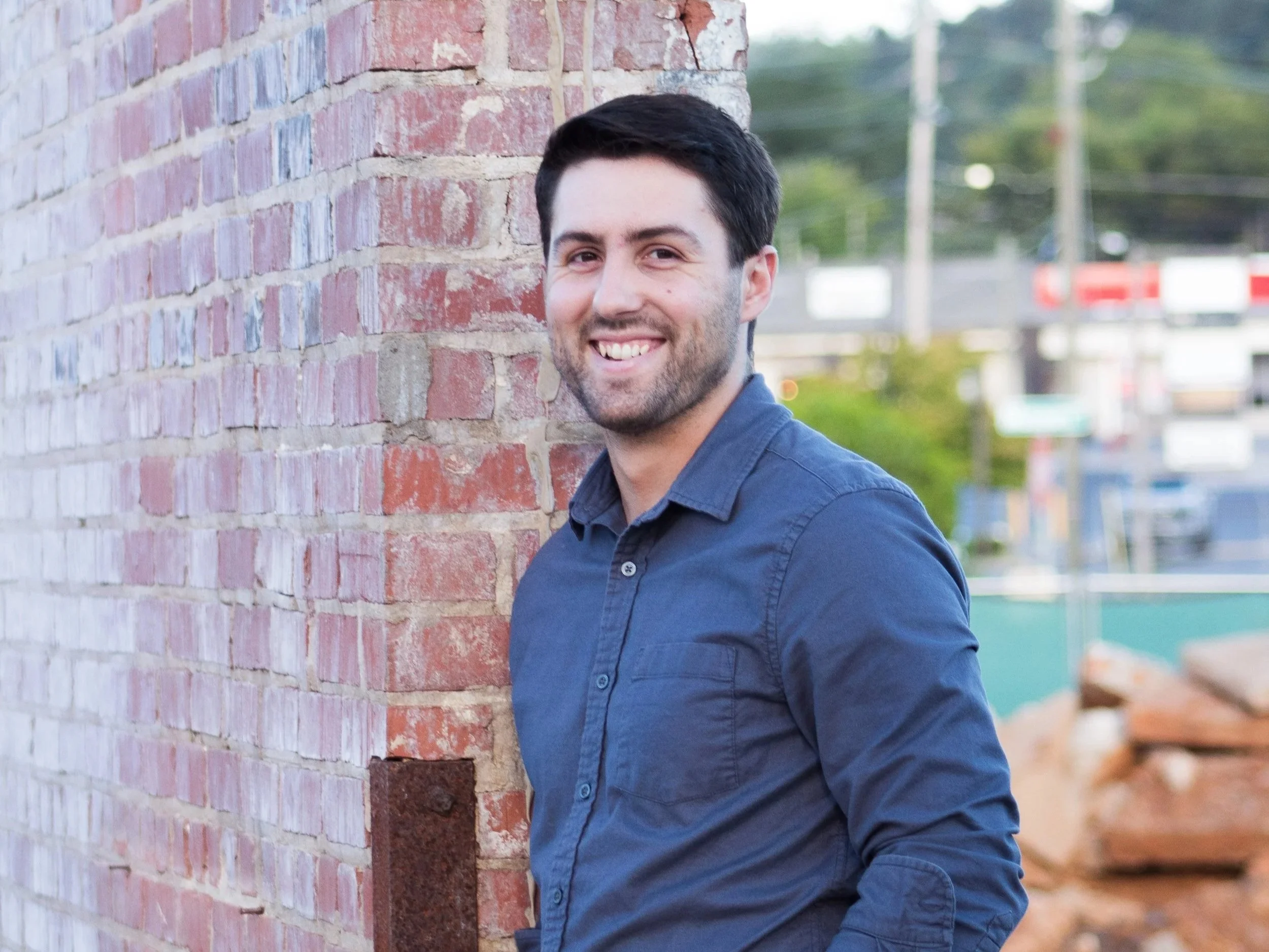 A headshot of Nick Larkins with dark hair and a beard smiling and wearing a blue button-up shirt, standing next to a red brick wall outdoors.