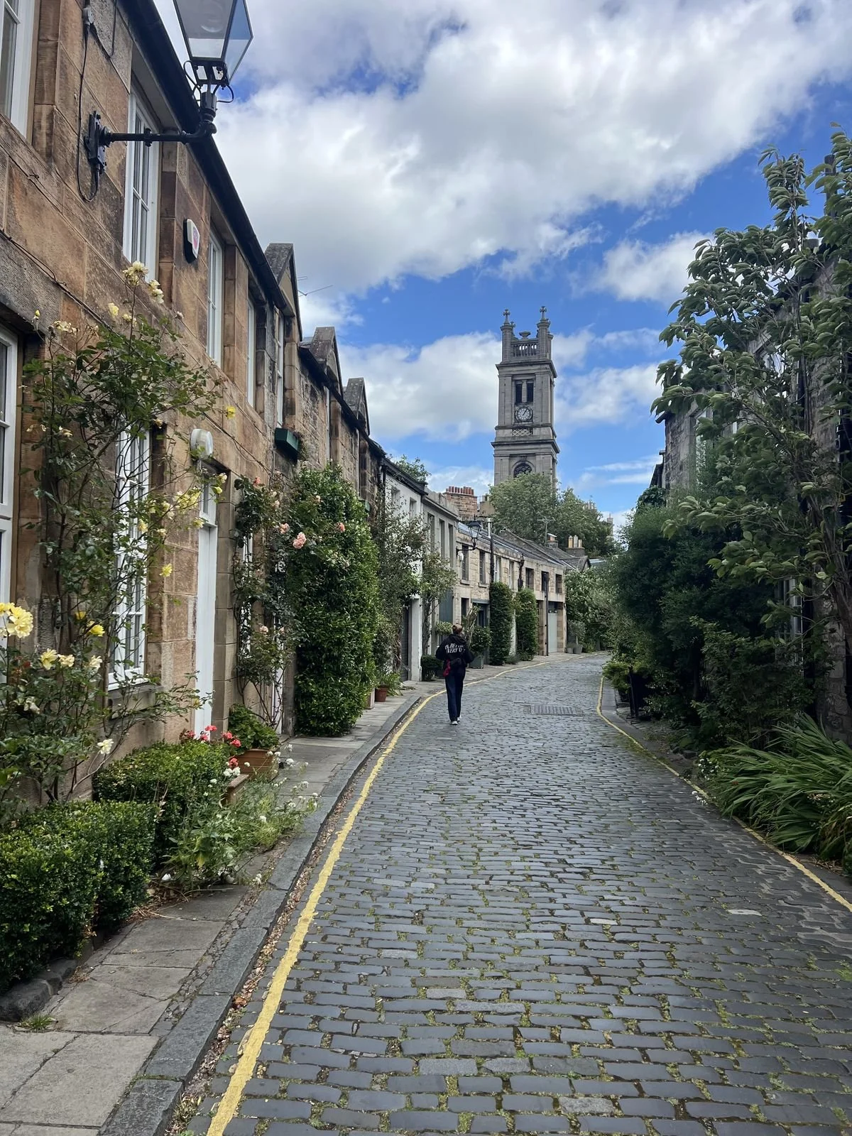 A cobblestone street lined with residential buildings and greenery, with a person walking away from the camera and a church tower in the background under a partly cloudy sky.