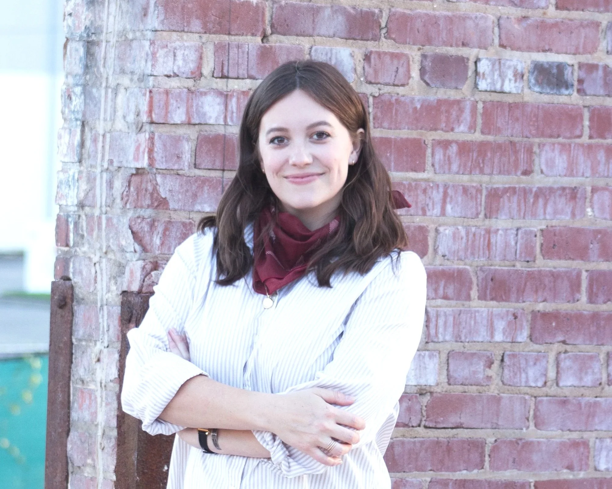 A headshot of Dannie Wright with brown hair smiling with arms crossed, standing against a brick wall.