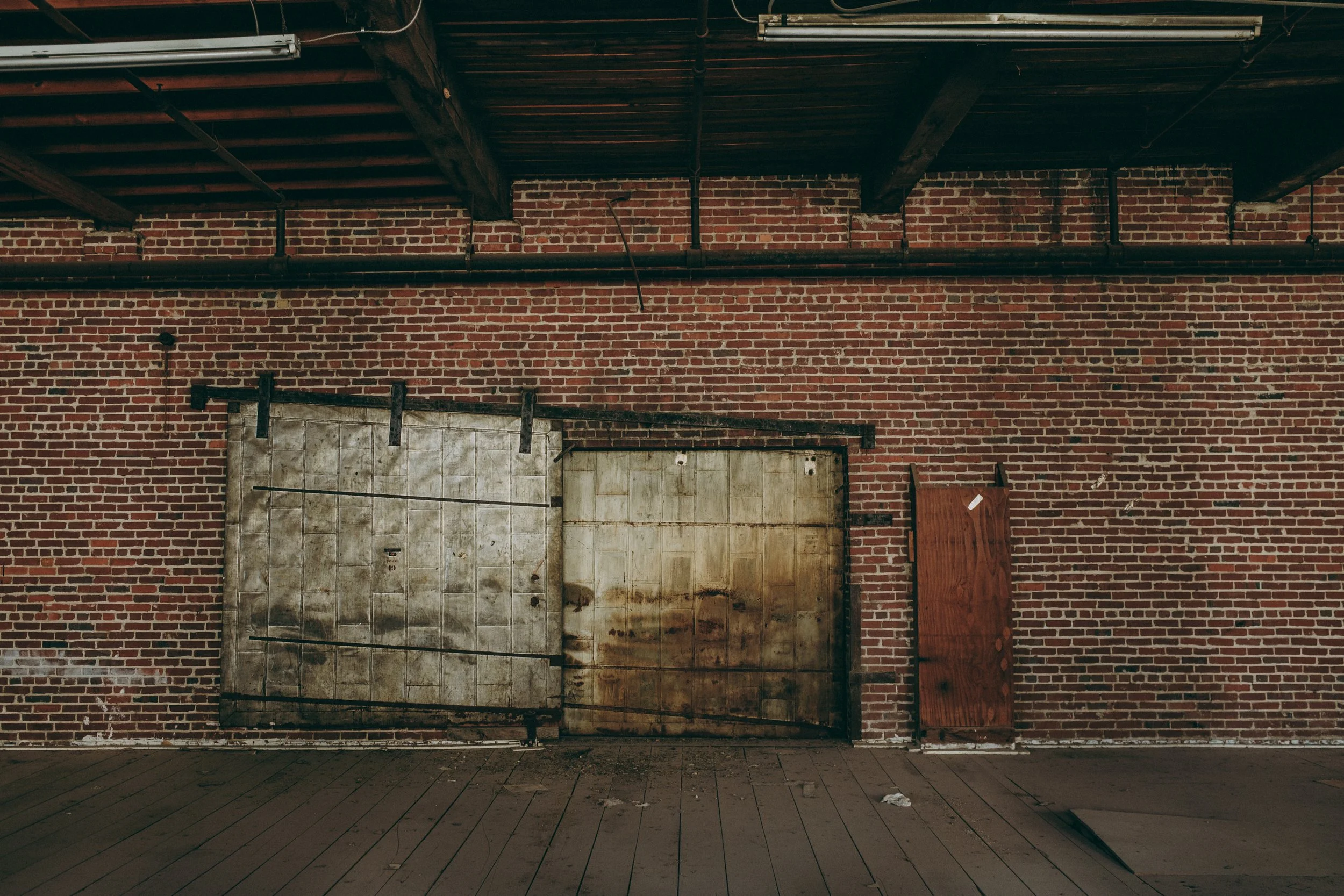 Interior of our main project's building with red brick walls, a wooden ceiling with black pipes, and a metal door with a padlock.