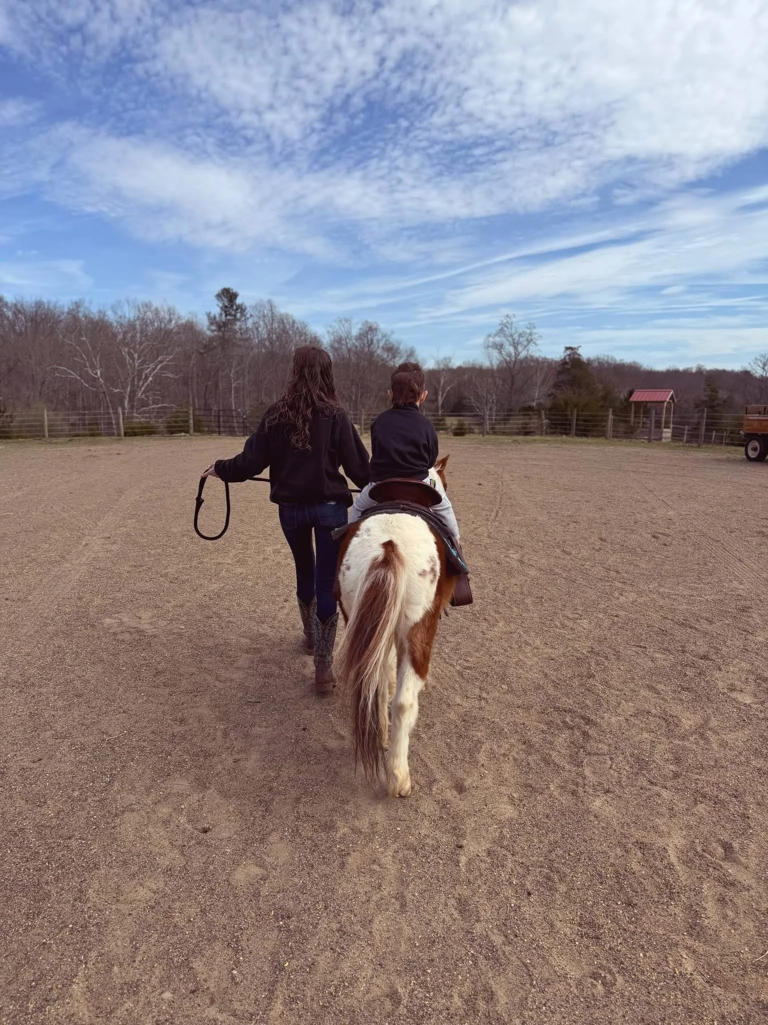 A person walking a child on horseback in an open dirt field with trees and a fence in the background under a blue sky with scattered clouds.