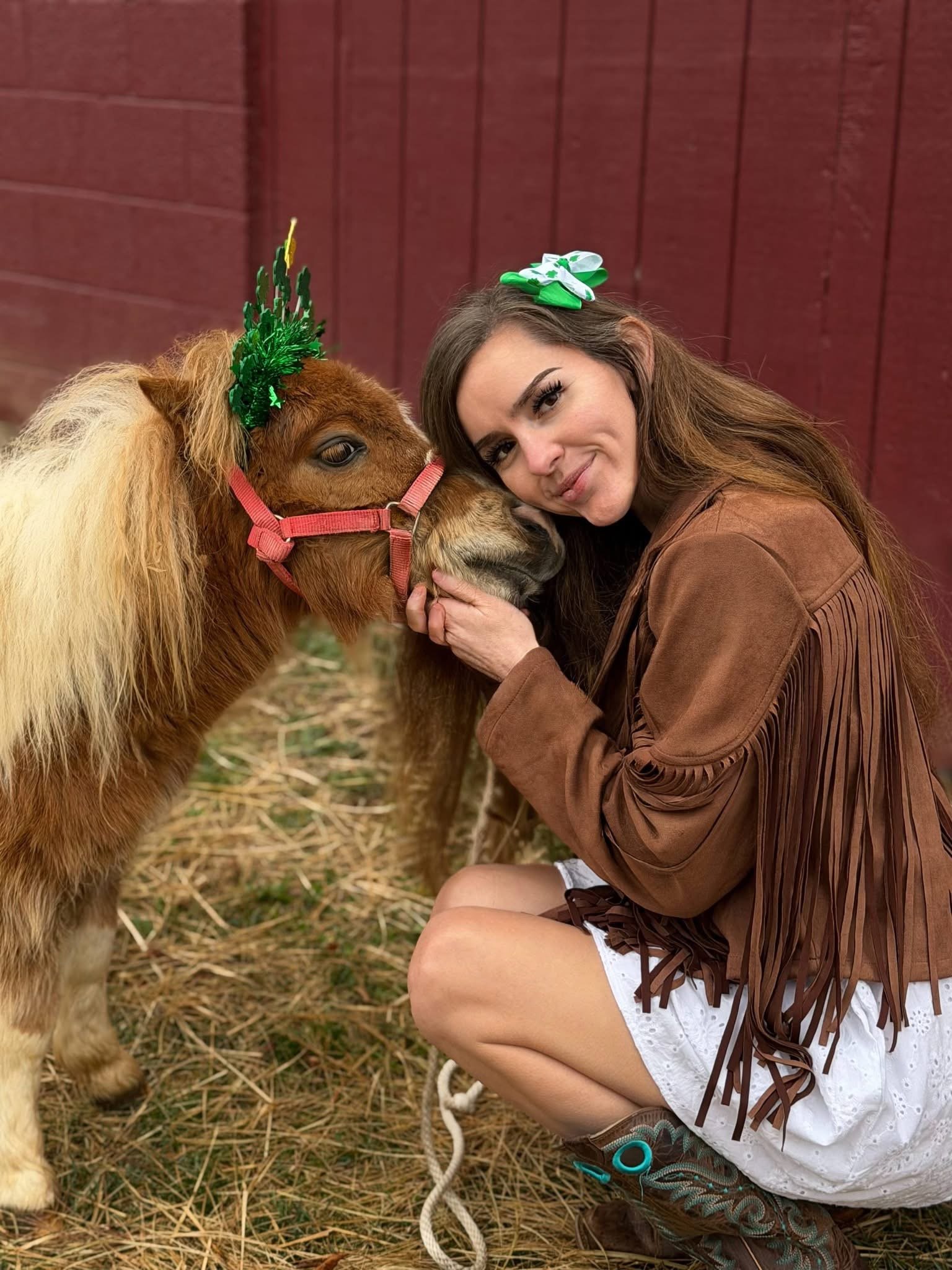 Woman in a brown fringed jacket and white dress crouching with a brown and white miniature horse, both wearing green holiday decorations on their heads, outdoors near a red wooden wall.