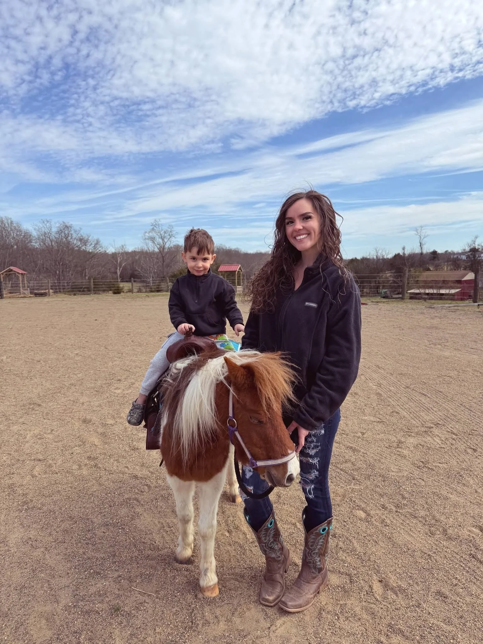 A woman and a young boy are outdoors, with the boy sitting on a pony and the woman standing beside it, both smiling. The background shows a fenced area with a cloudy blue sky.