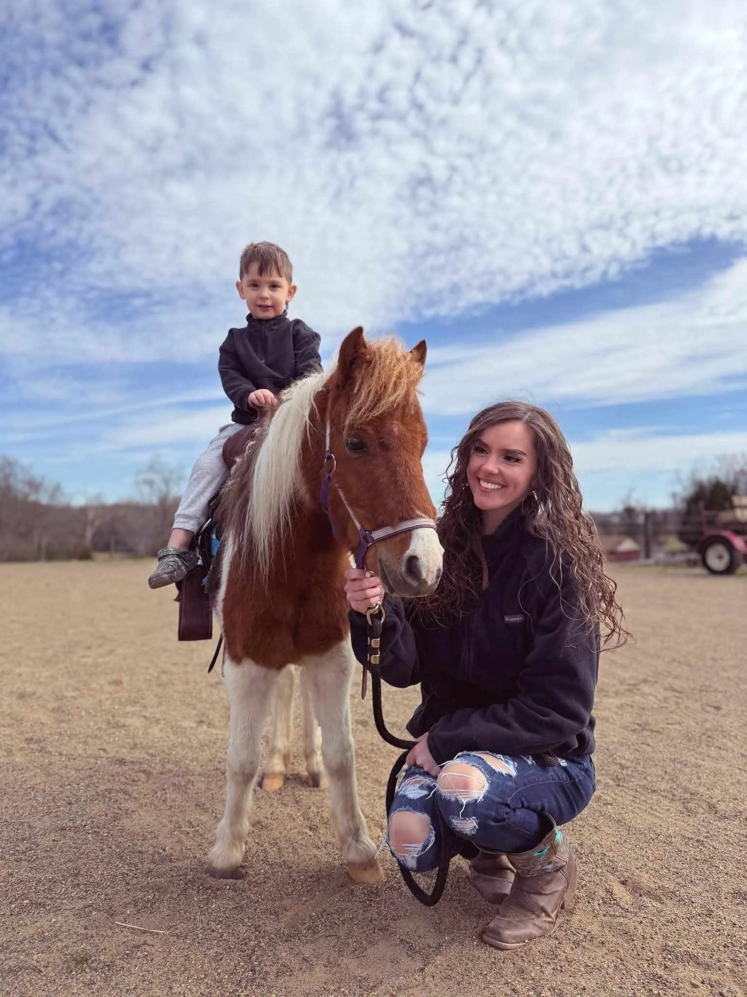 Woman kneeling beside a brown and white pony, with a young boy riding on its back against a blue sky with clouds.