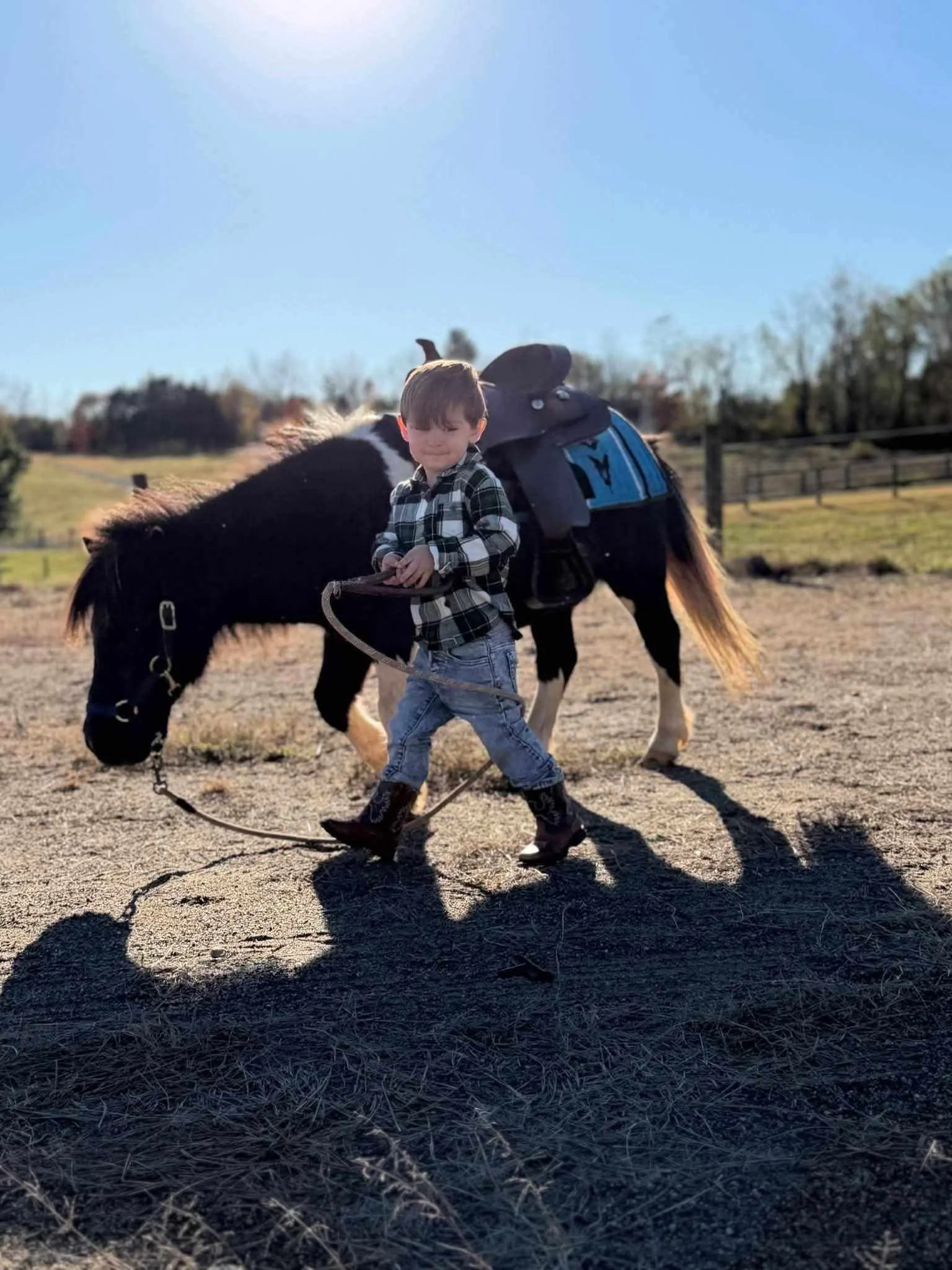 A young boy walking on a farm with a pony, holding its lead rope, and a saddle on its back.