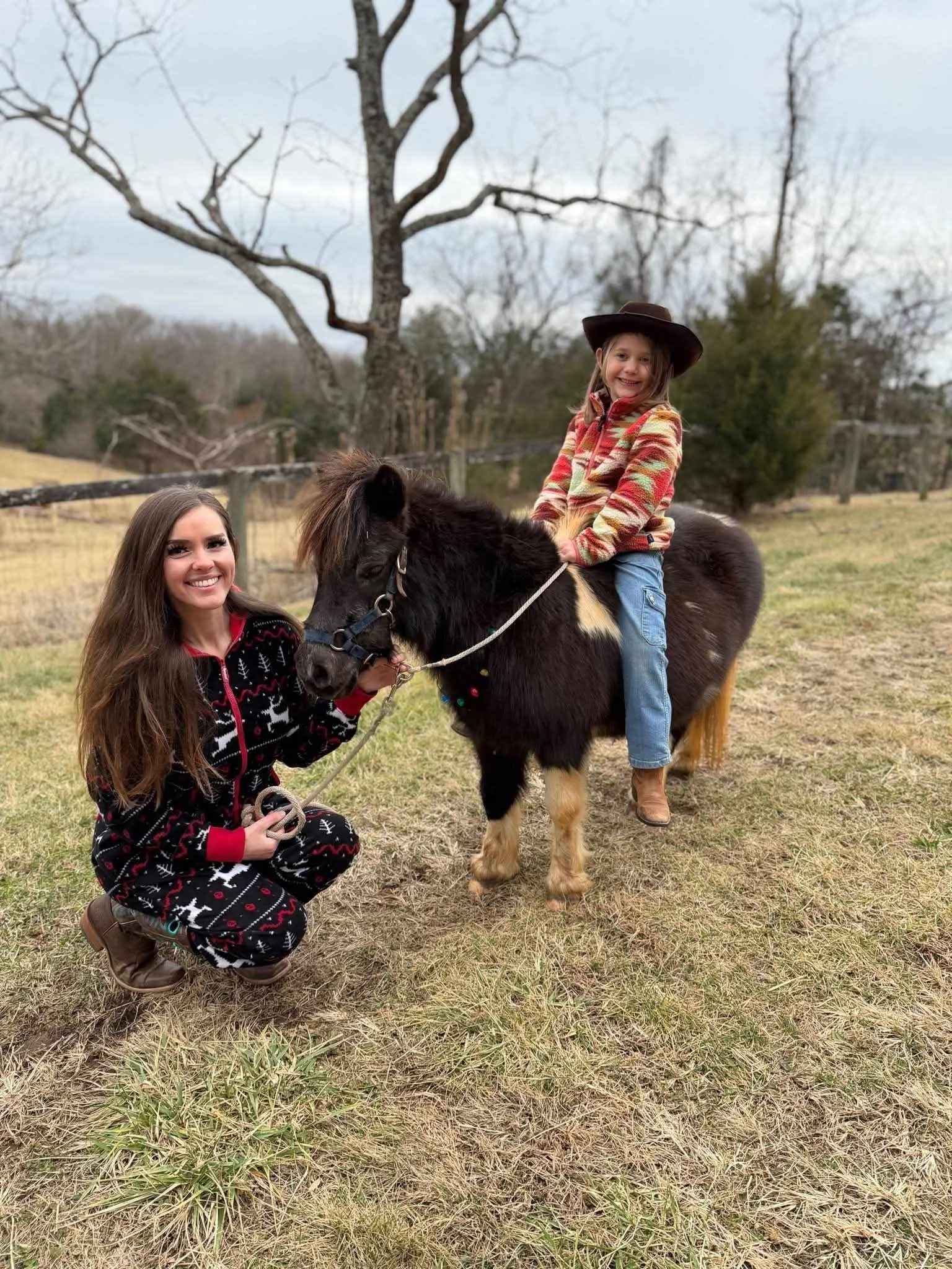 Young girl sitting on a small pony horse in a grassy outdoor setting, with a woman kneeling beside the horse, holding the reins and smiling at the camera. The girl is wearing a western hat and colorful jacket, and the woman is dressed in holiday-them