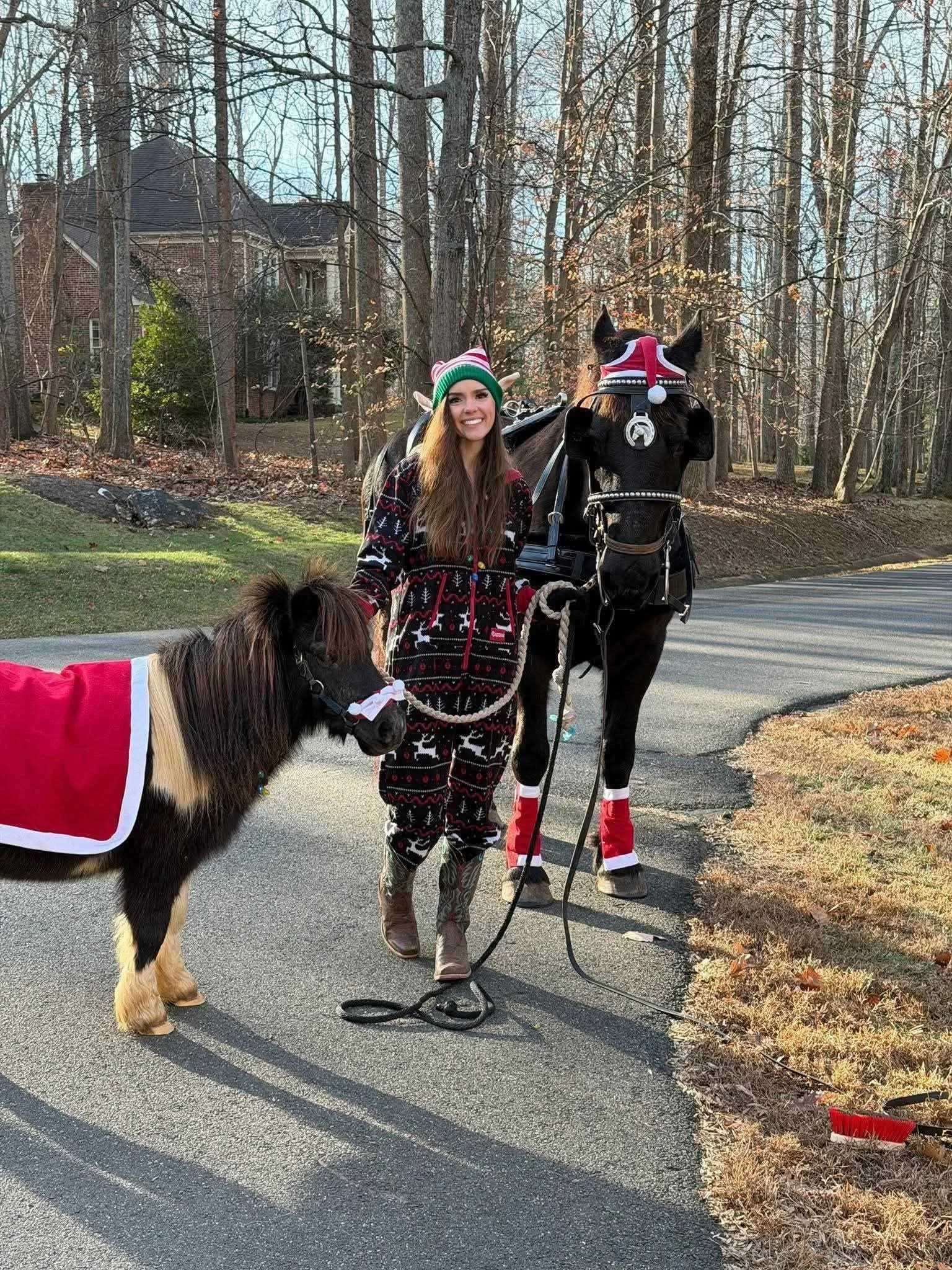 A young woman dressed in Christmas-themed pajamas and a festive hat stands on a paved road holding the lead of a small dog in a red holiday cape. Next to her, a black horse wearing a Santa hat, red leg wraps, and a bridle stands in a wooded area with