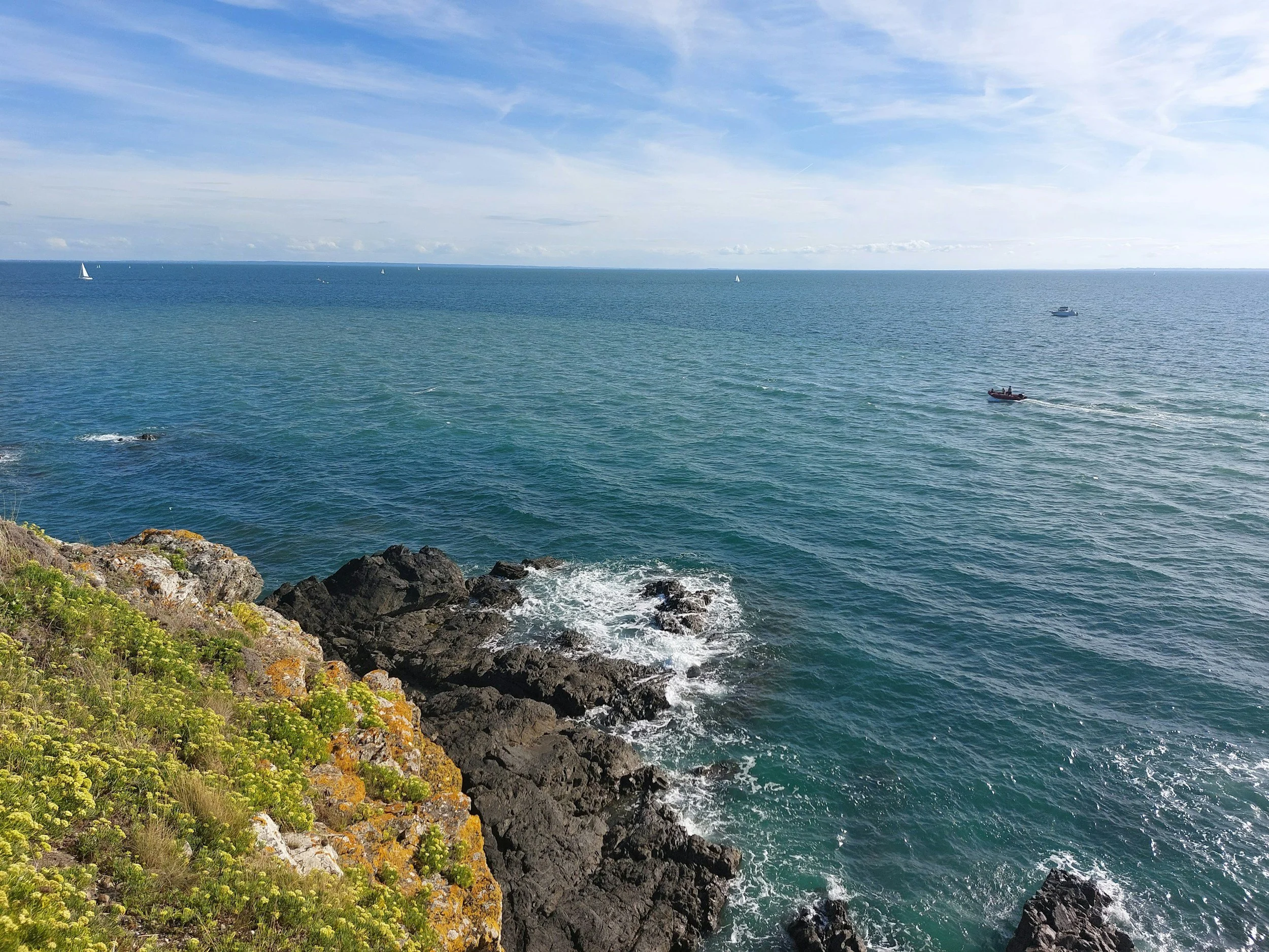 A scenic view of the ocean with several boats sailing, rocky shoreline with green plants, and a partly cloudy blue sky.