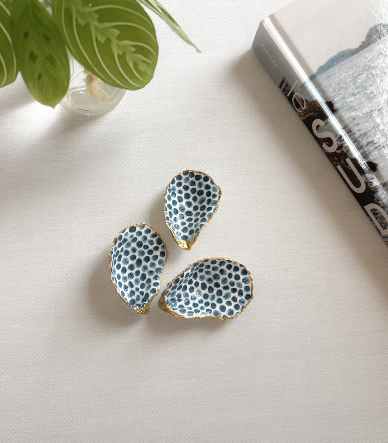Three ceramic dishes with blue polka dot pattern and gold edges, placed on a white surface, next to a green potted plant and a book.