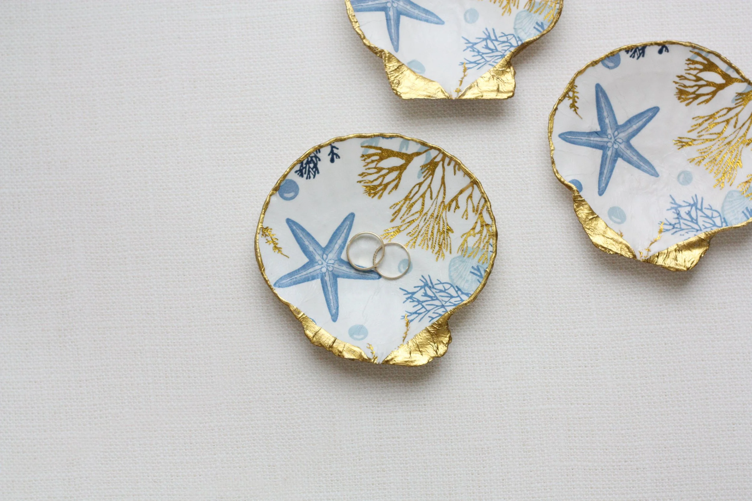Three ceramic dishes with gold-edged rims, decorated with blue starfish, seashells, and coral patterns, arranged on a textured beige background. Two simple ring bands are placed inside the bottom dish.