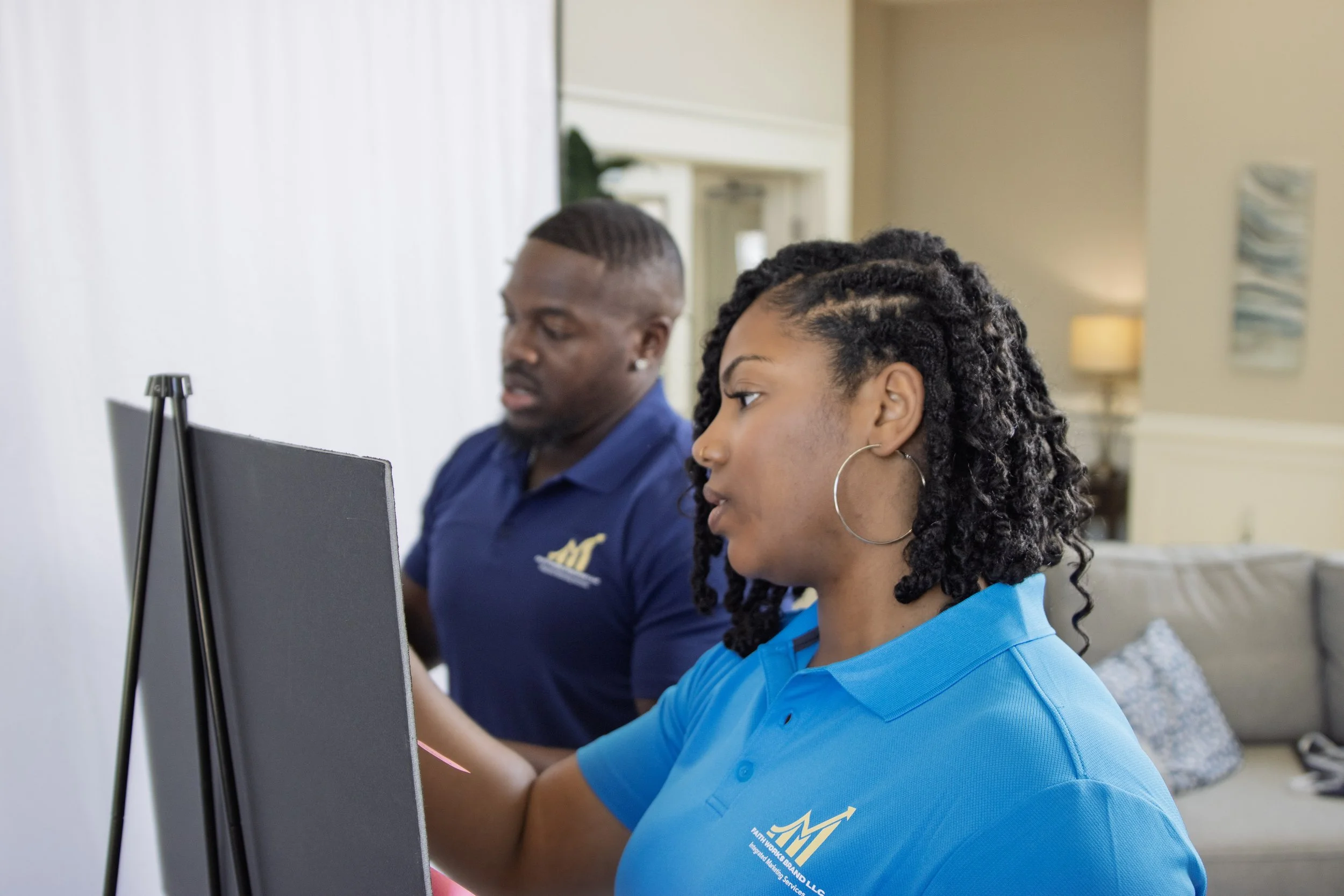 A woman with curly hair wearing a blue shirt with a logo on it, focusing on a computer screen, with a man in the background also looking at a computer, in an office setting.