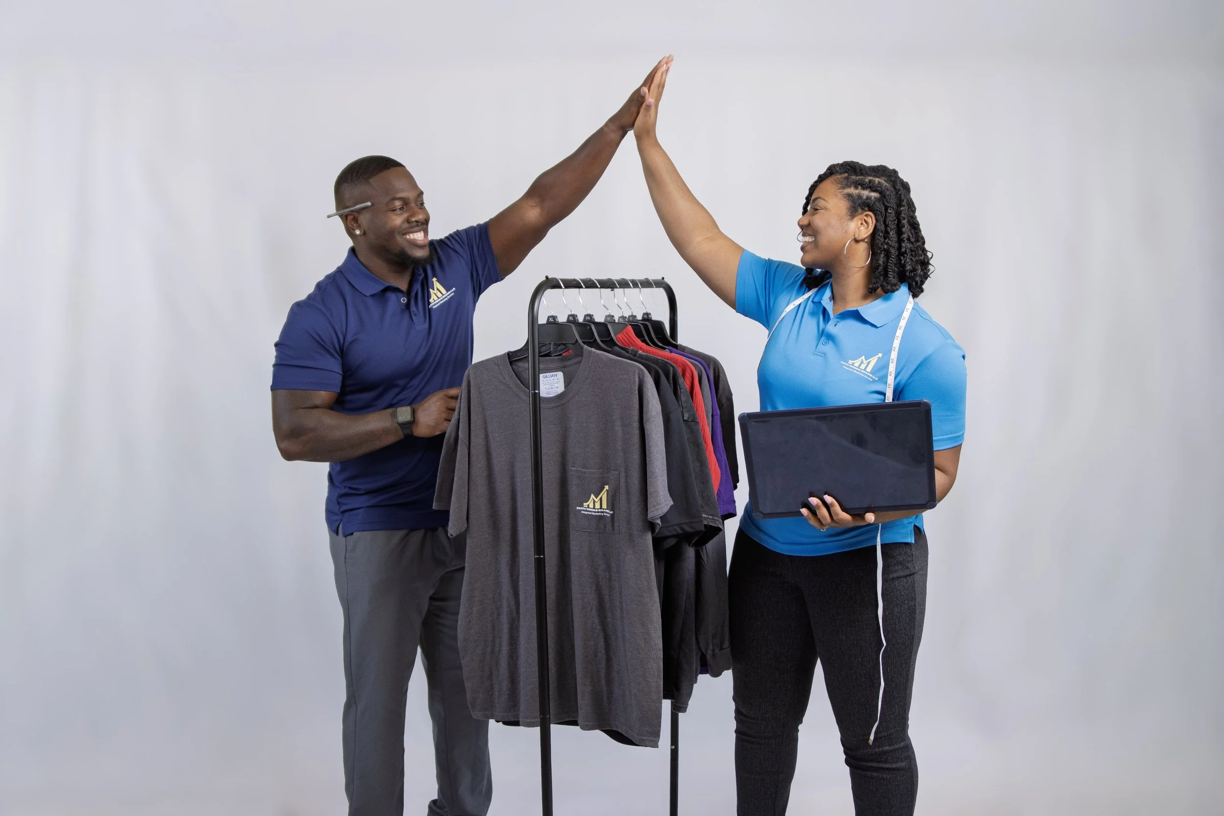 A man and a woman high-fiving each other in a retail or promotional setting. The man is wearing a blue polo shirt with a logo, and the woman is wearing a blue polo shirt with a measuring tape around her neck. They are standing behind a clothing rack with various shirts and a laptop on a table.