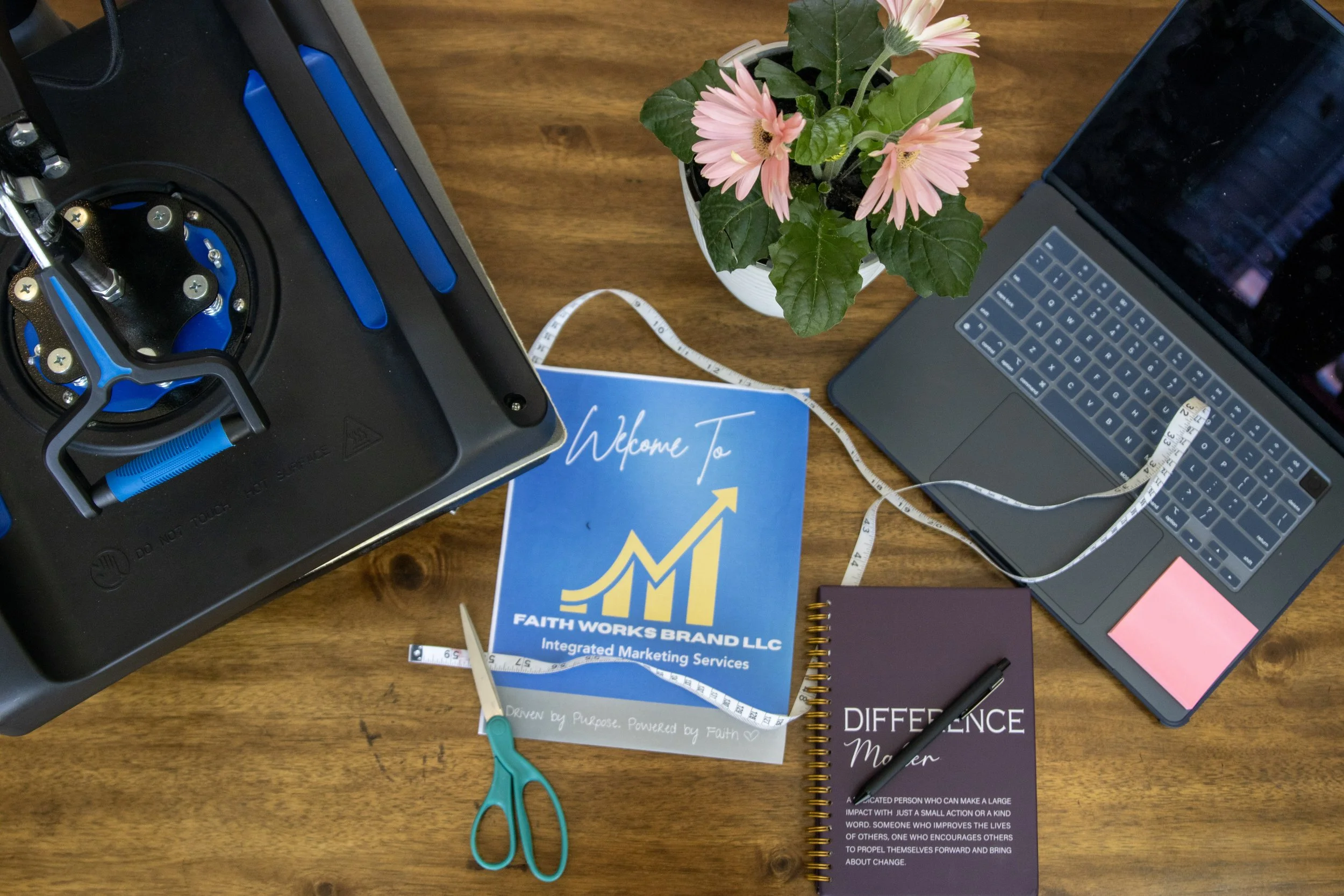 Top-down view of a wooden desk with a computer monitor, a silver laptop, a potted pink daisy plant, a blue booklet titled 'Faith Works Brand LLC,' a small purple notebook, a black pen, a pair of blue scissors, tape measure, and a sticky note.