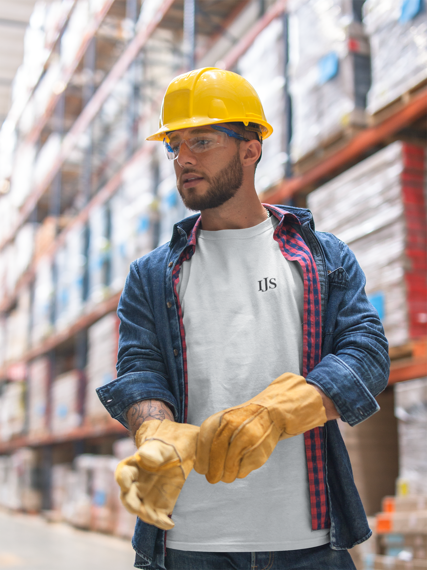 A man wearing a yellow safety helmet, safety glasses, a white t-shirt with 'IJS' on it, and a denim jacket with red plaid lining, handling a pair of yellow work gloves in a warehouse with shelves filled with boxes.