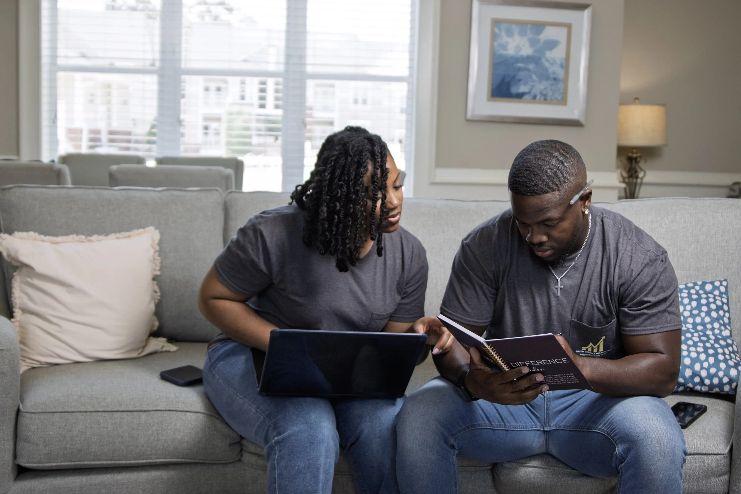 A man and a woman sitting on a gray couch, looking at a magazine together in a living room with large windows, a framed picture on the wall, a lamp, and pillows.