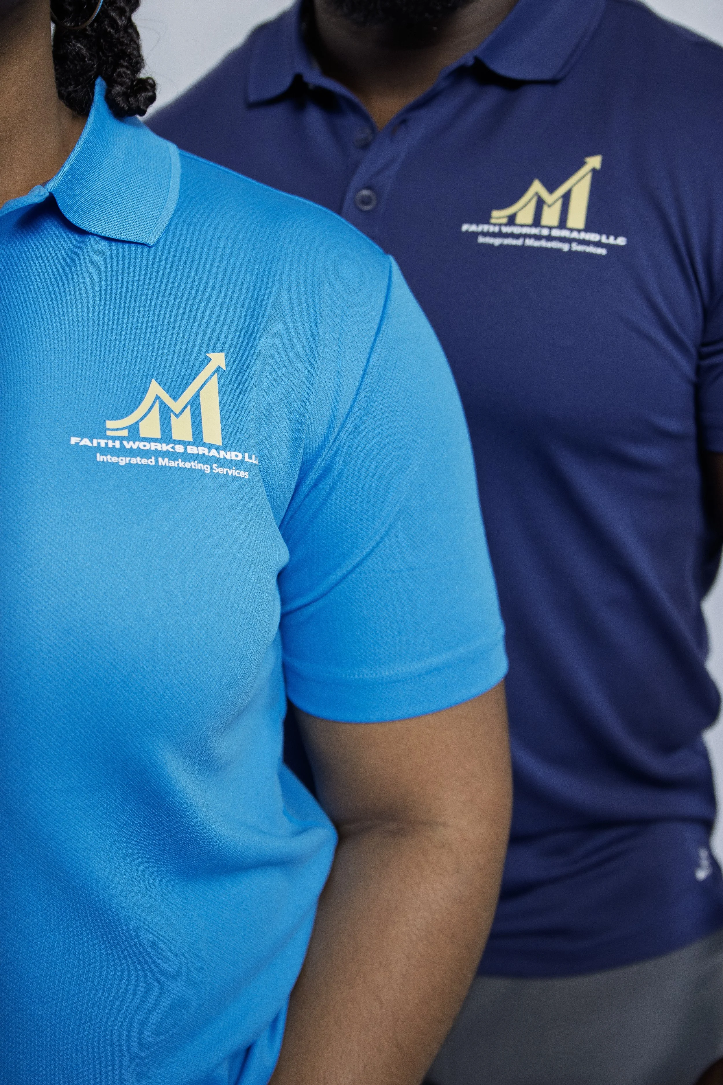 Close-up of two people wearing polo shirts with a logo that reads 'Faith Works Brand LLC' and 'Integrated Marketing Services' against a gray background.
