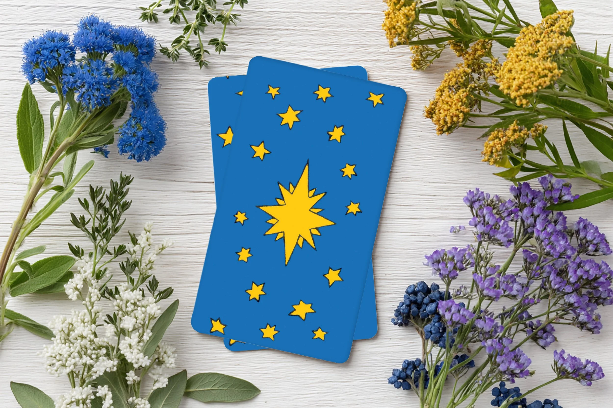 Des cartes de tarot avec un symbole d'étoile brillante jaune sur un fond bleu, entourées de fleurs colorées telles que bleu, blanc, jaune et violet sur une surface en bois blanc.
