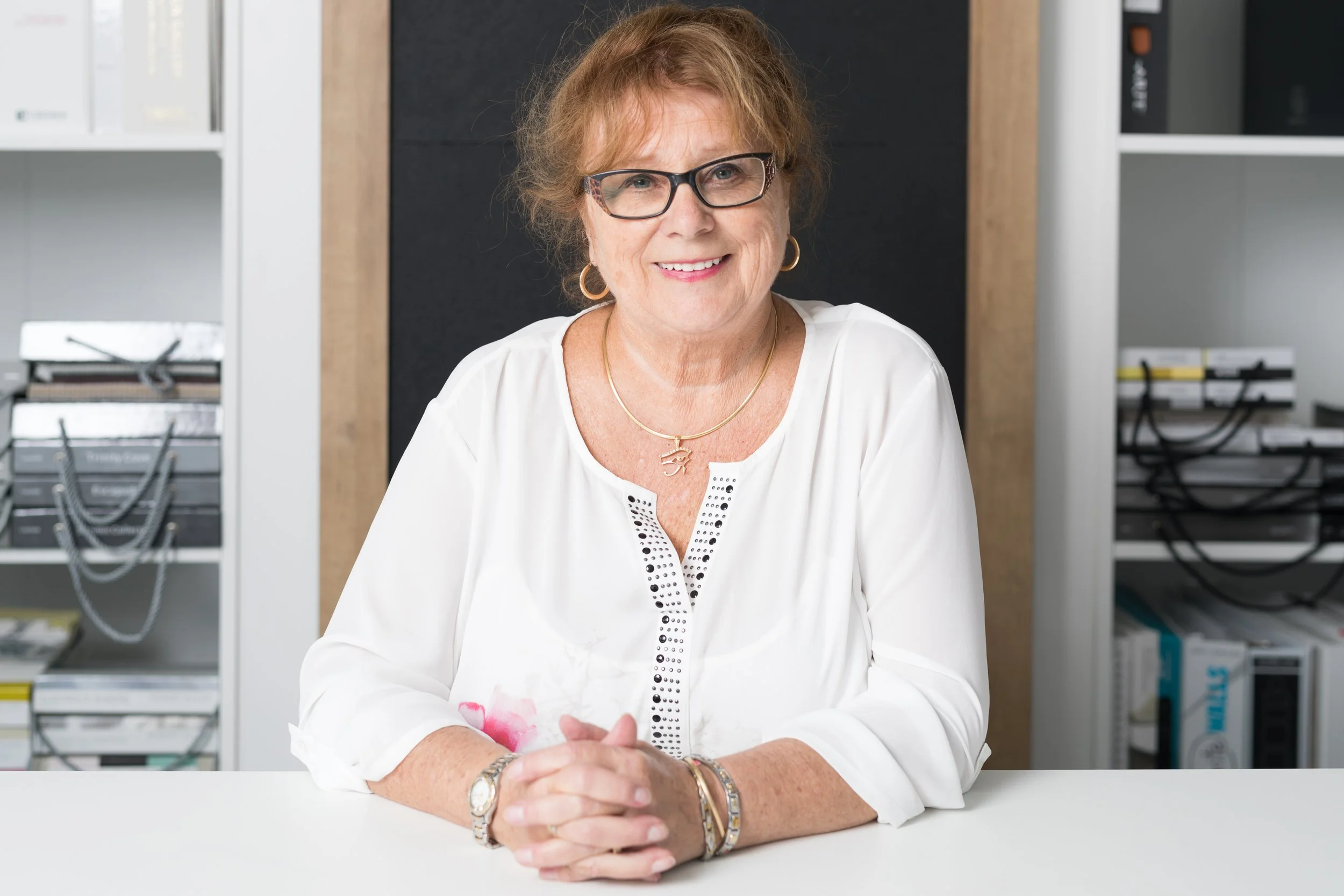 Une femme âgée, souriante, portant des lunettes et une chemise blanche, assise à une table dans un bureau