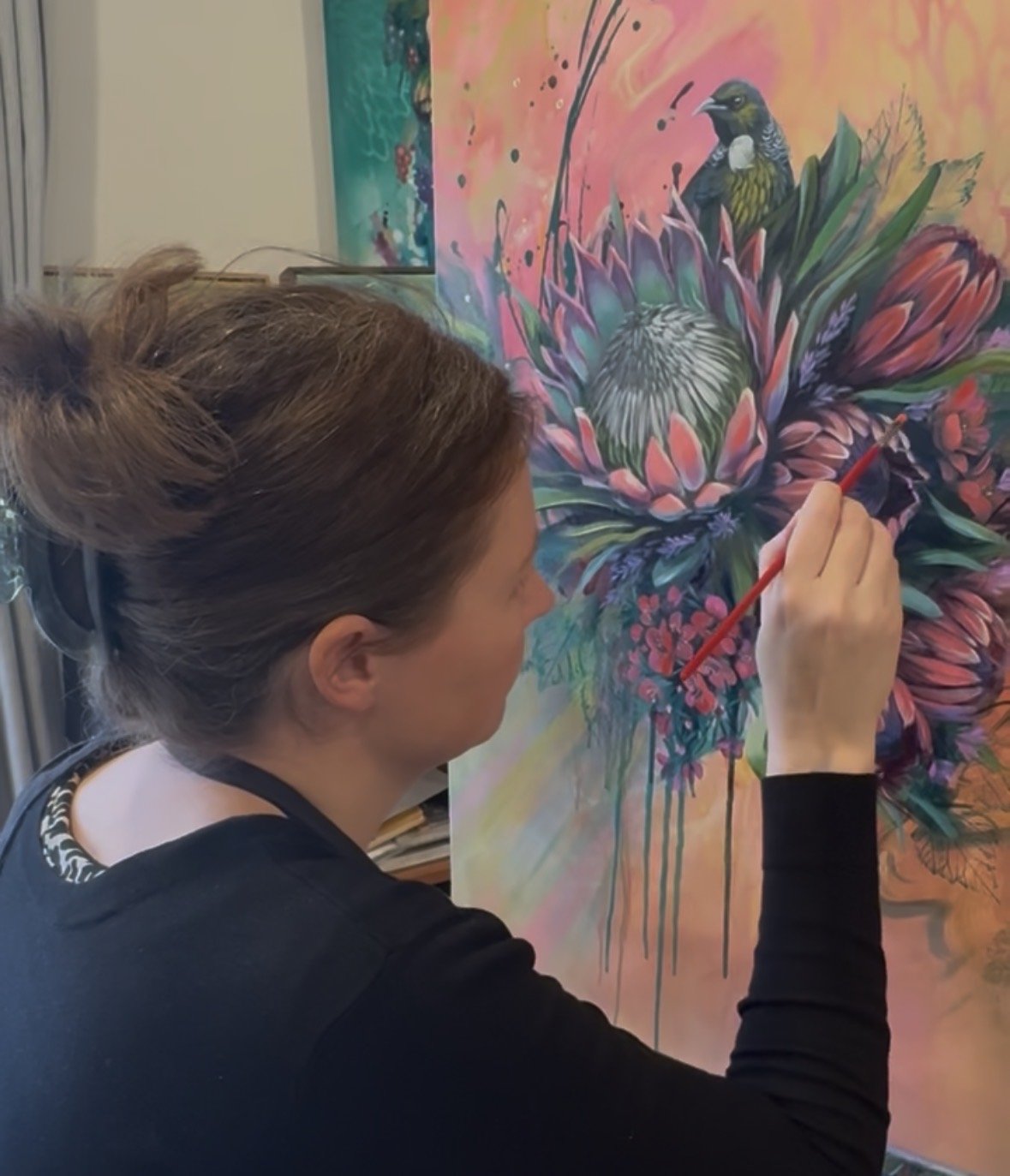 New Zealand artist Jessica Jane Holliday adding the finishing details to a colourful native bird and floral painting in her studio.