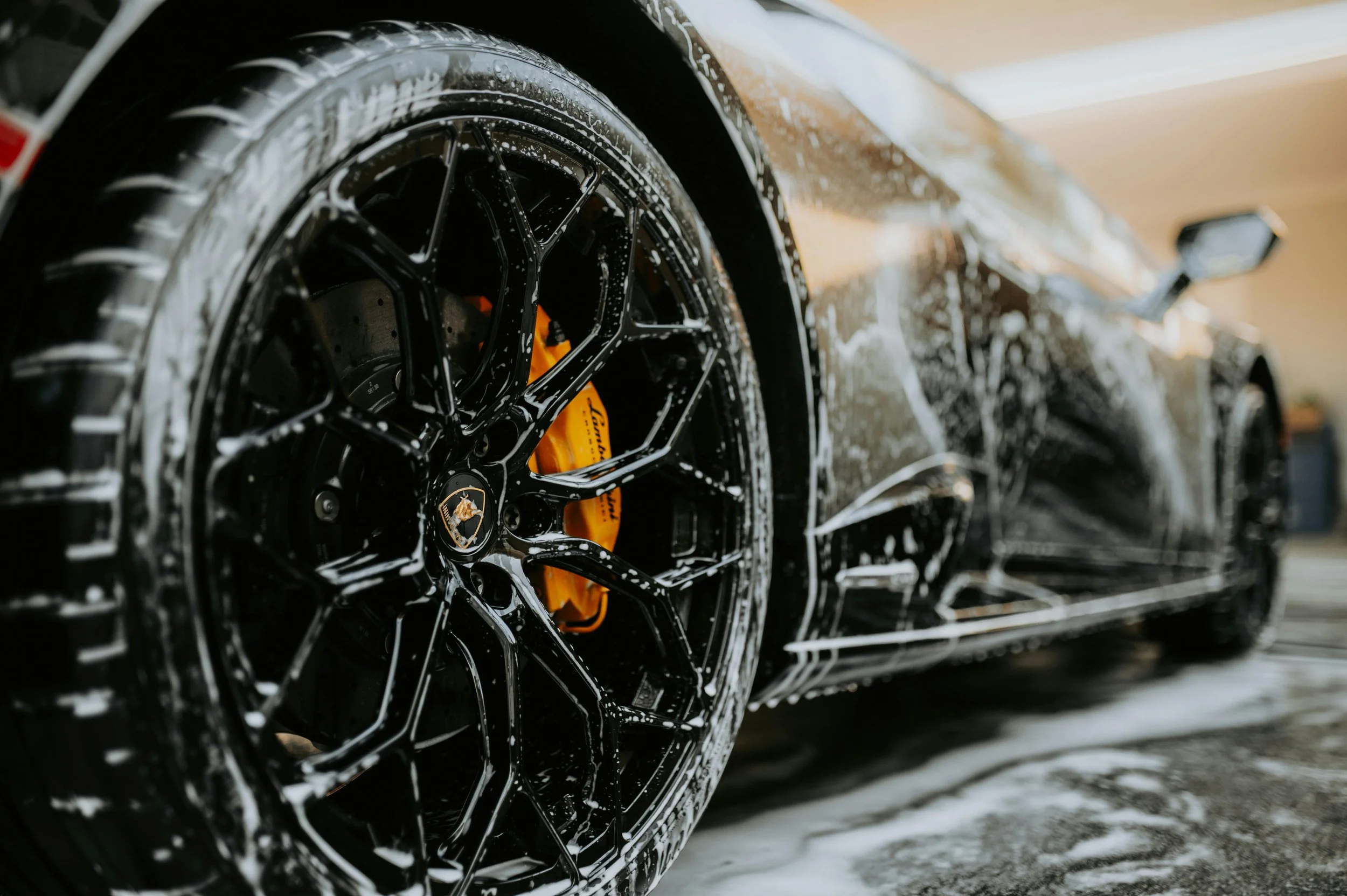 Close-up of a black luxury sports car's wheel covered in soap suds, highlighting the gold brake caliper.