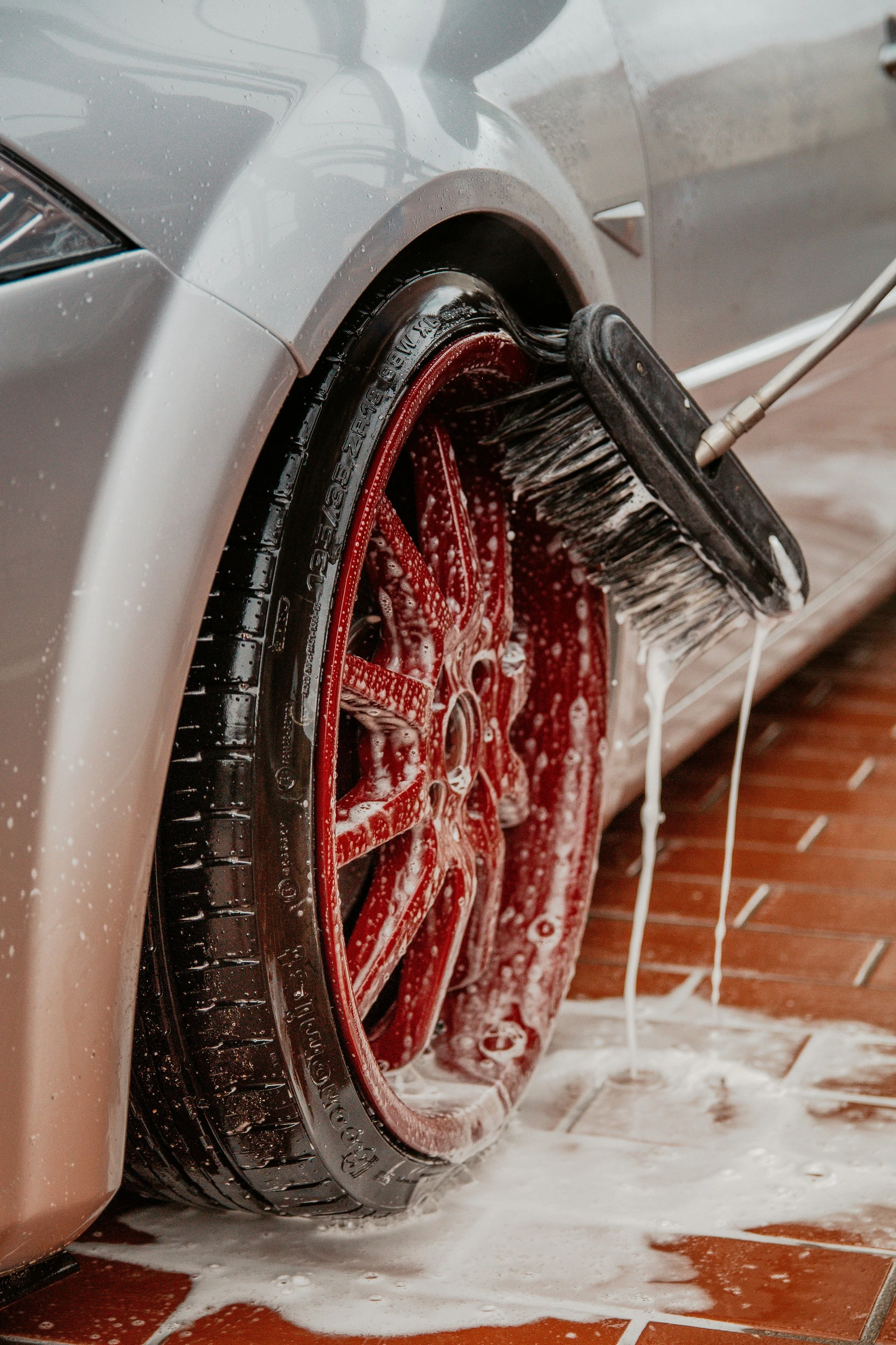 Close-up of a silver car wheel being cleaned with a brush, soap suds overflowing onto a tiled floor.