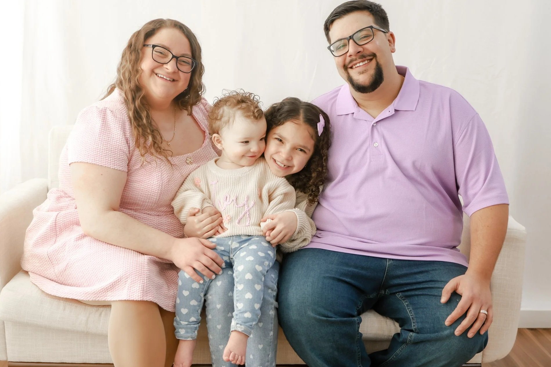 A family of four sitting on a beige couch with a white background, smiling and embracing each other. The family includes a woman with glasses and long glasses, a man with glasses and a beard, a young girl with curly hair and a purple bow, and a toddler girl with curly red hair.