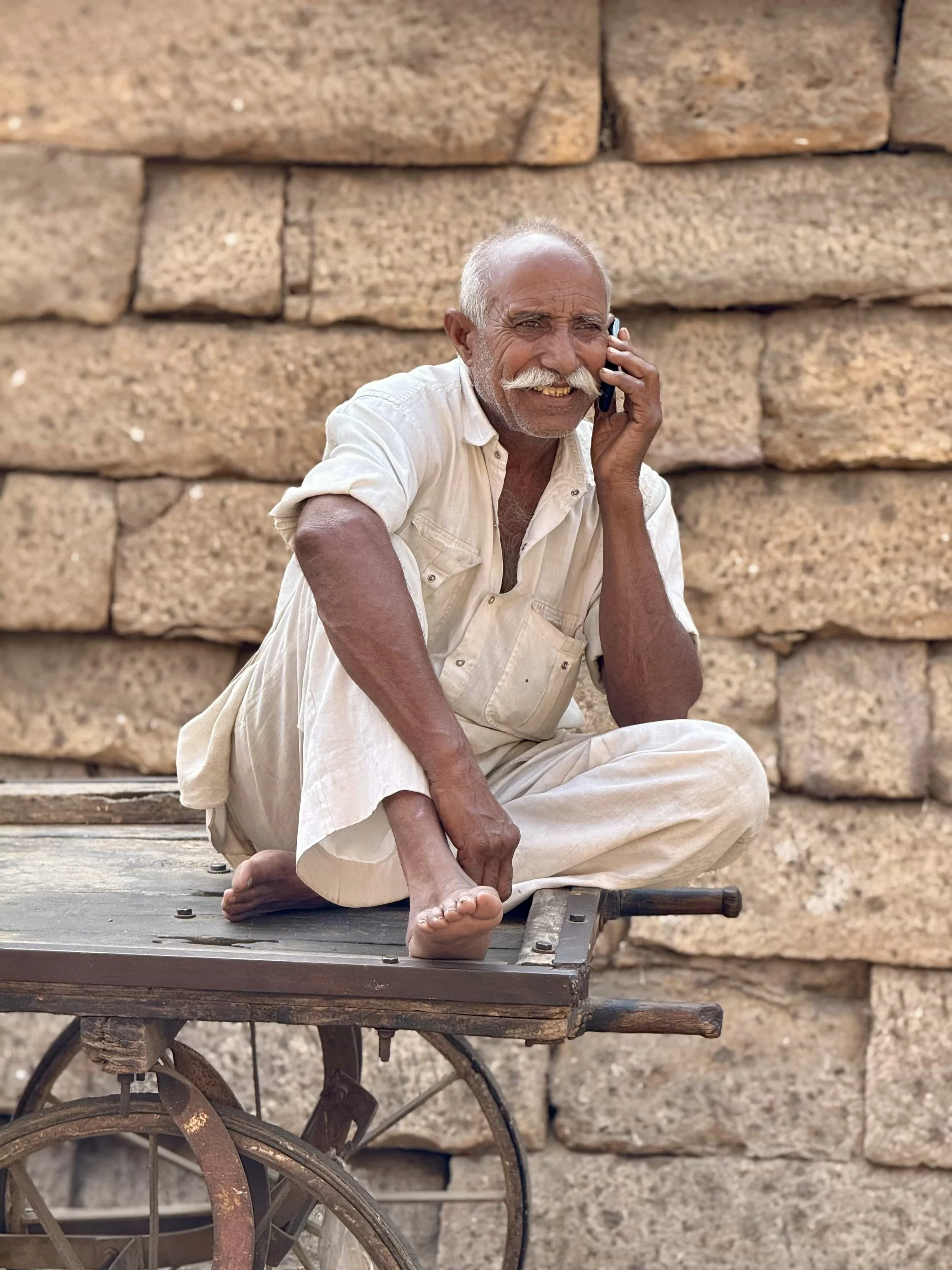 An elderly man with gray hair and mustache sitting on a wooden cart, talking on a cell phone, with a brick wall in the background.