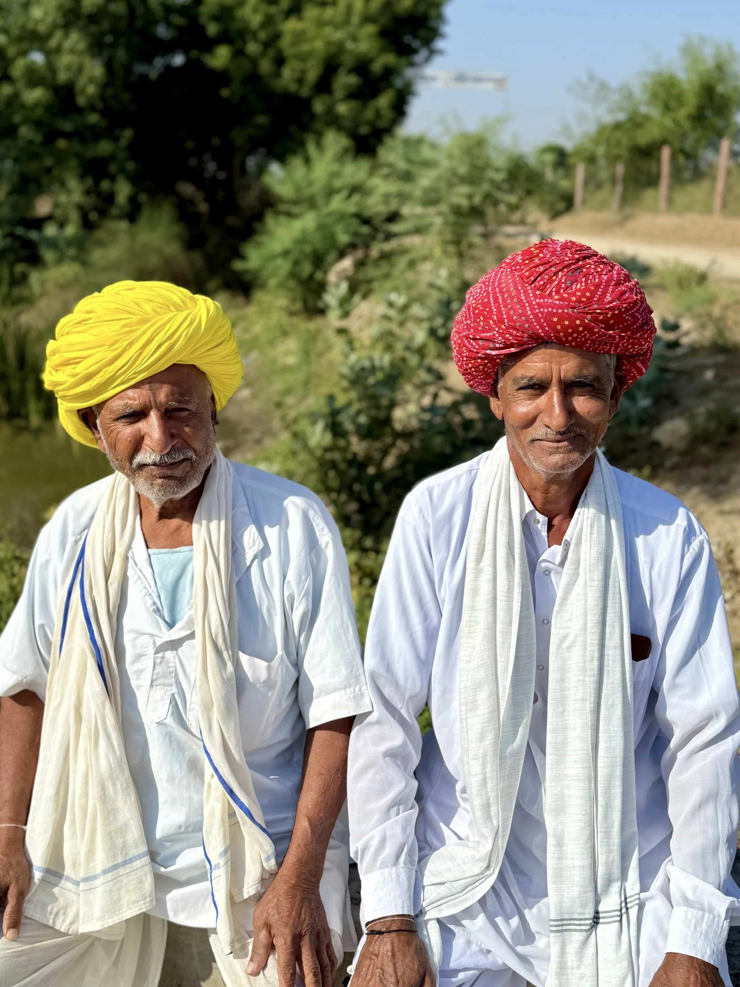 Two elderly men wearing traditional Indian attire with turbans, standing outdoors in a rural setting, holding hands.