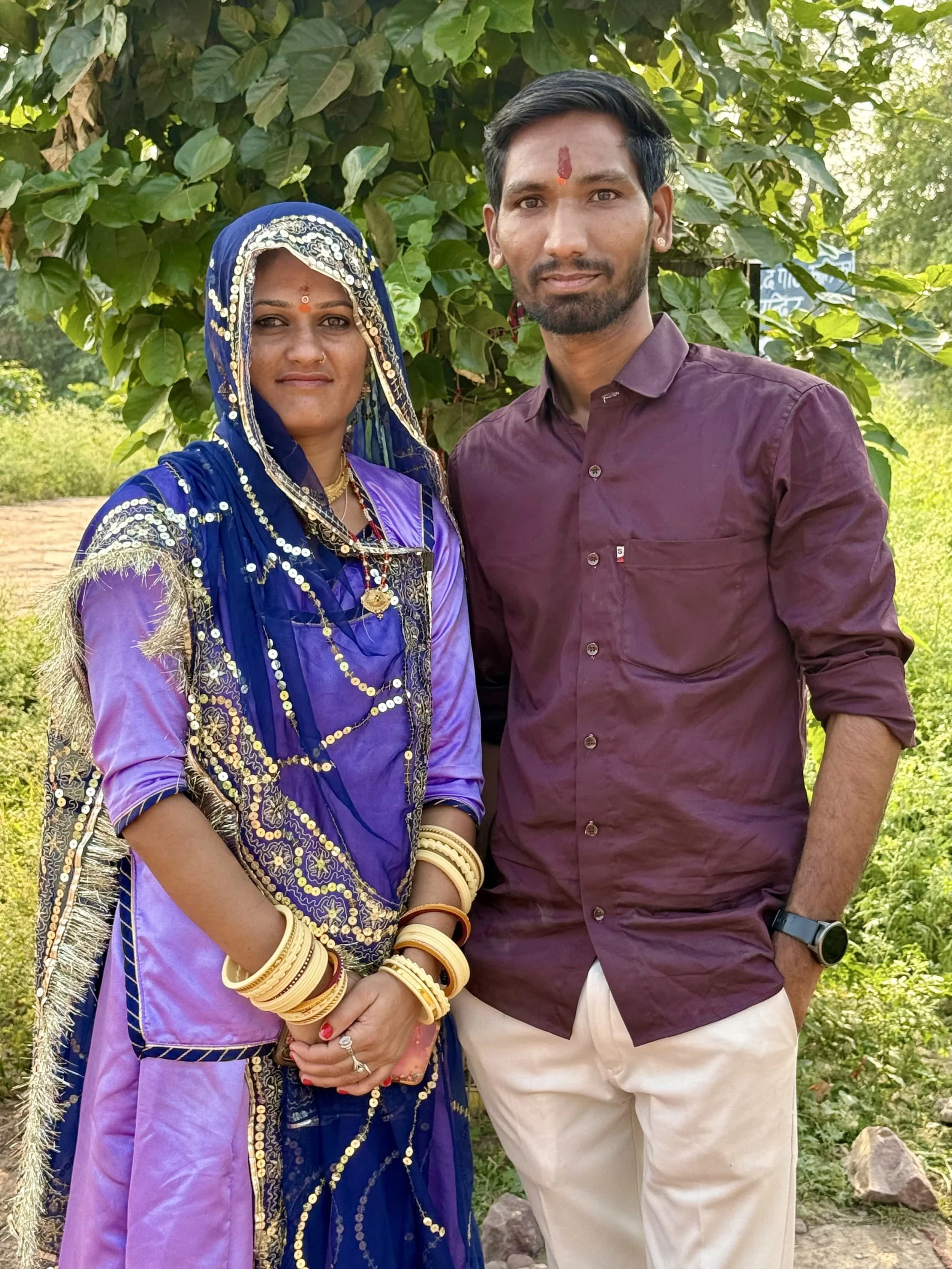 A man and a woman standing outdoors in front of green foliage, dressed in traditional Indian clothing. The woman is wearing a purple saree with gold embroidery and jewelry, and the man is wearing a maroon shirt and beige pants.