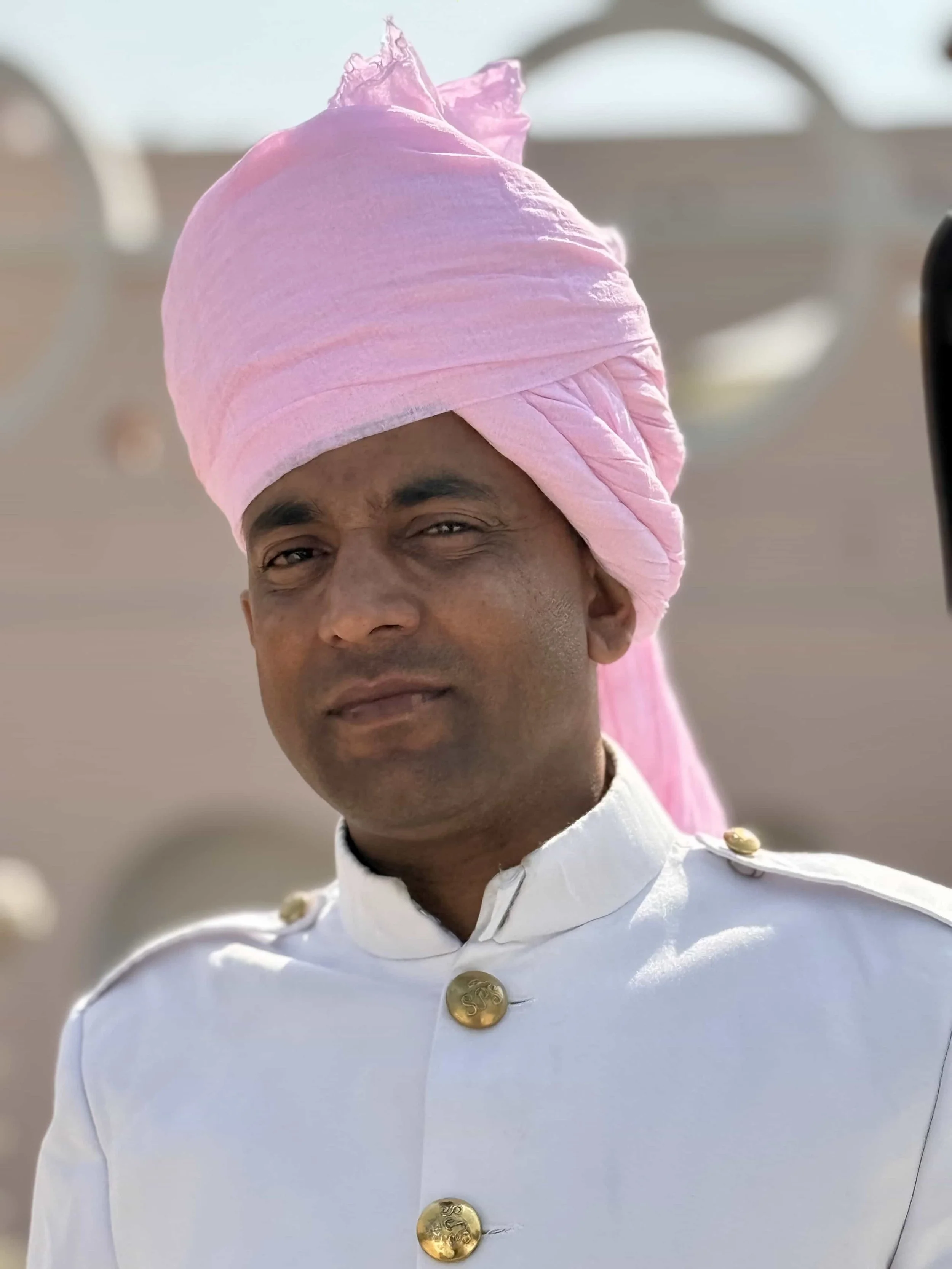 A man wearing a light pink turban and a white military uniform with gold buttons.