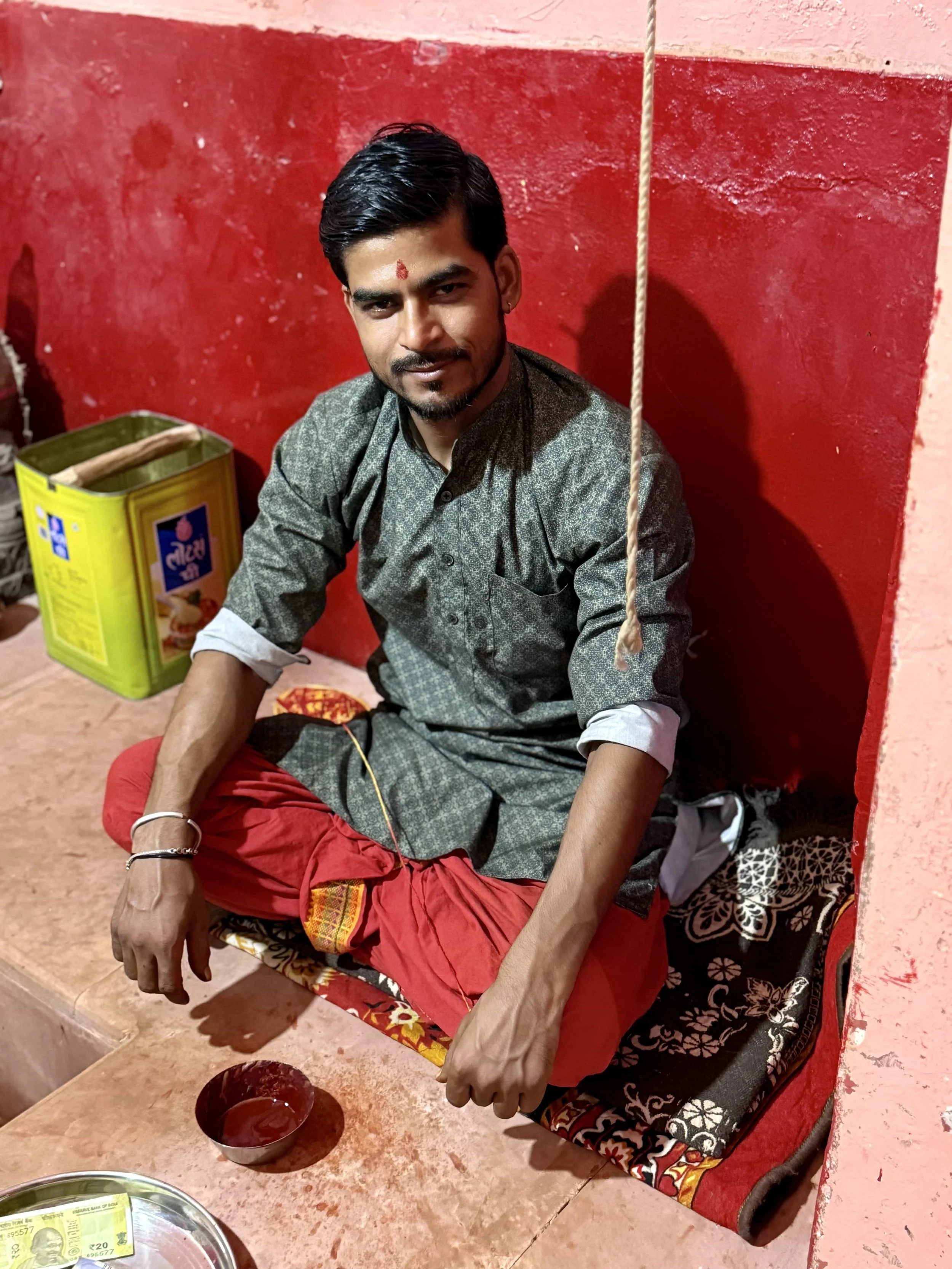 A young man with dark hair and a moustache sitting on a red cushioned seat against a red painted wall. He is wearing traditional clothing and has a small red mark on his forehead. There are two bowls in front of him, one with red liquid. There is als