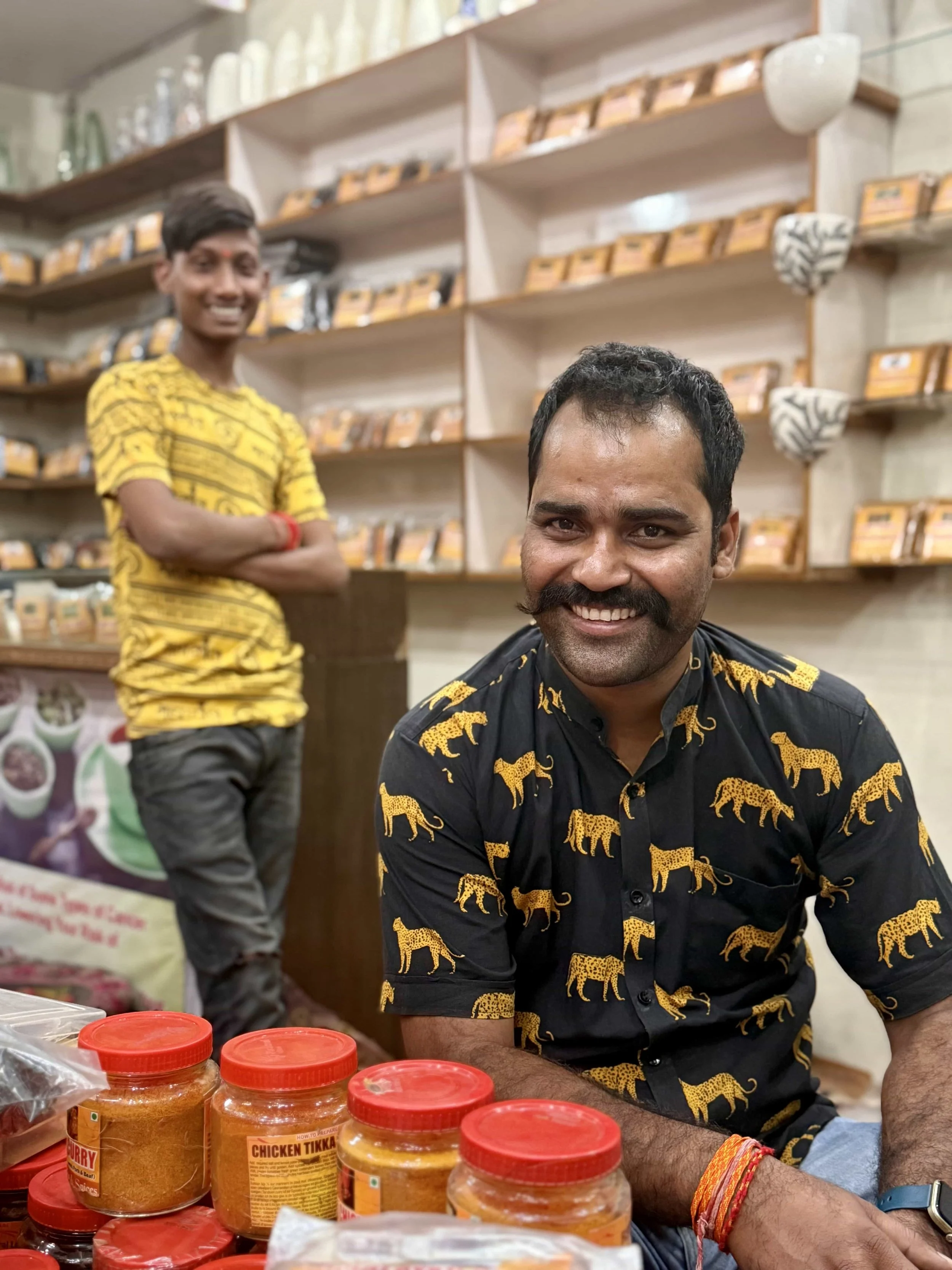 Two smiling men in a shop, one sitting in the foreground and the other standing in the background. The man in the foreground has short black hair, a beard, and is wearing a black shirt with yellow tiger patterns. The man in the background has short d