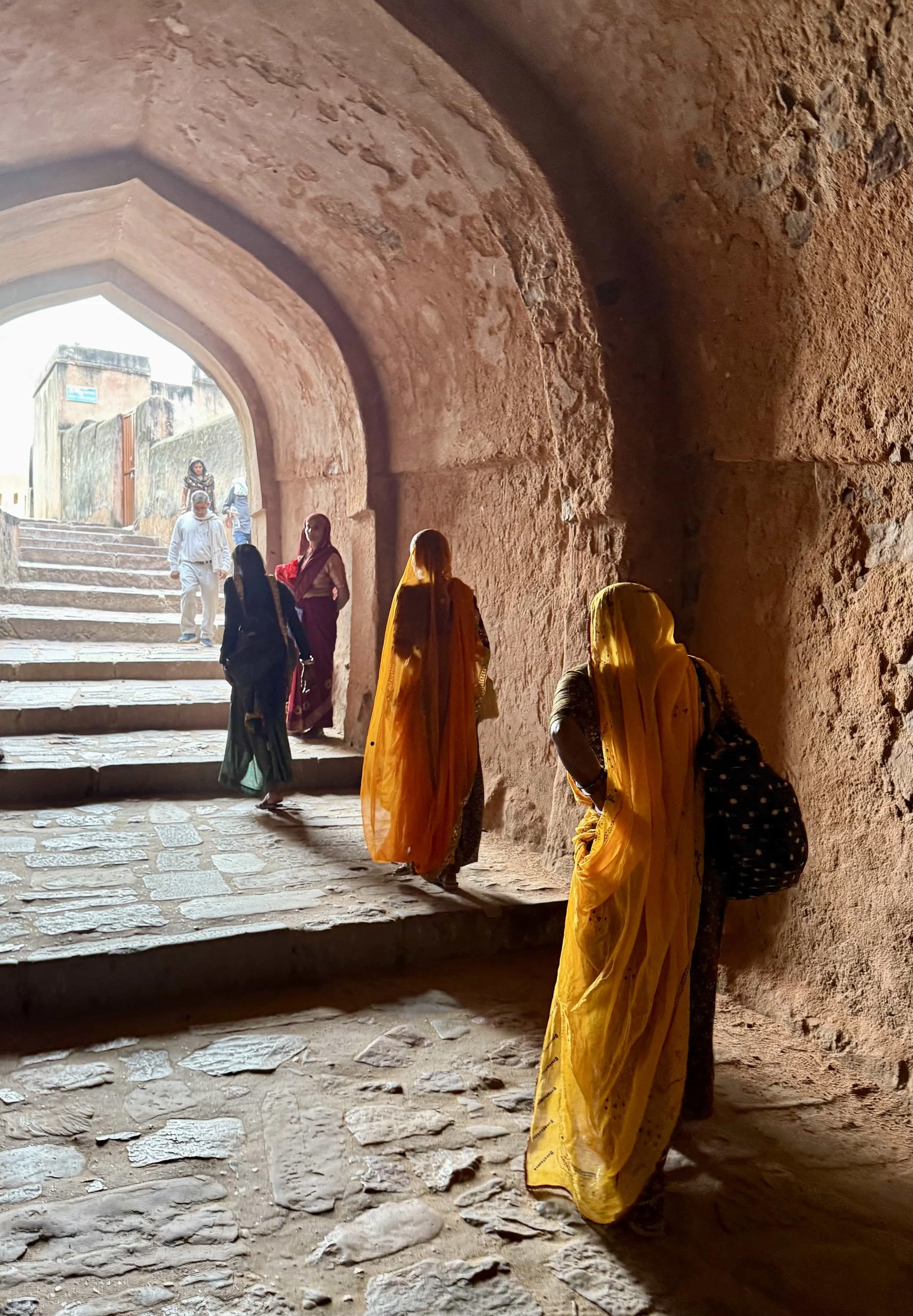 Women and children walking through a stone archway in a historic site, some wearing traditional clothing, with stone steps leading outside.