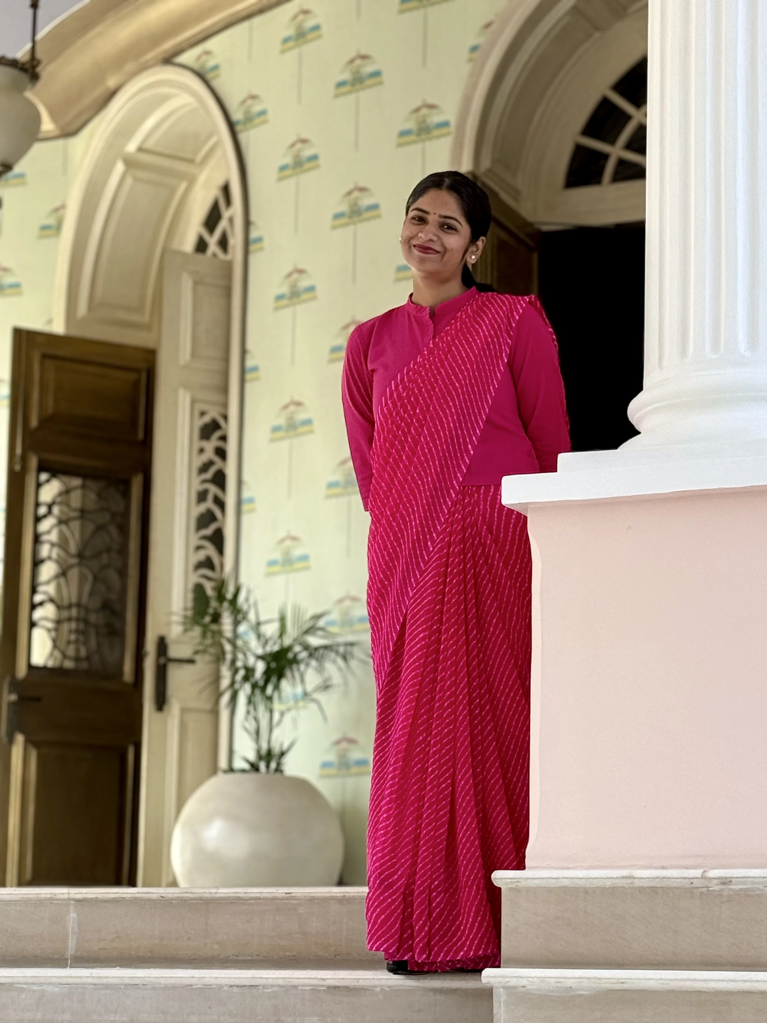 A woman wearing a bright pink traditional Indian outfit, standing on a staircase in an elegant interior with arches, a column, a potted plant, and patterned wallpaper.