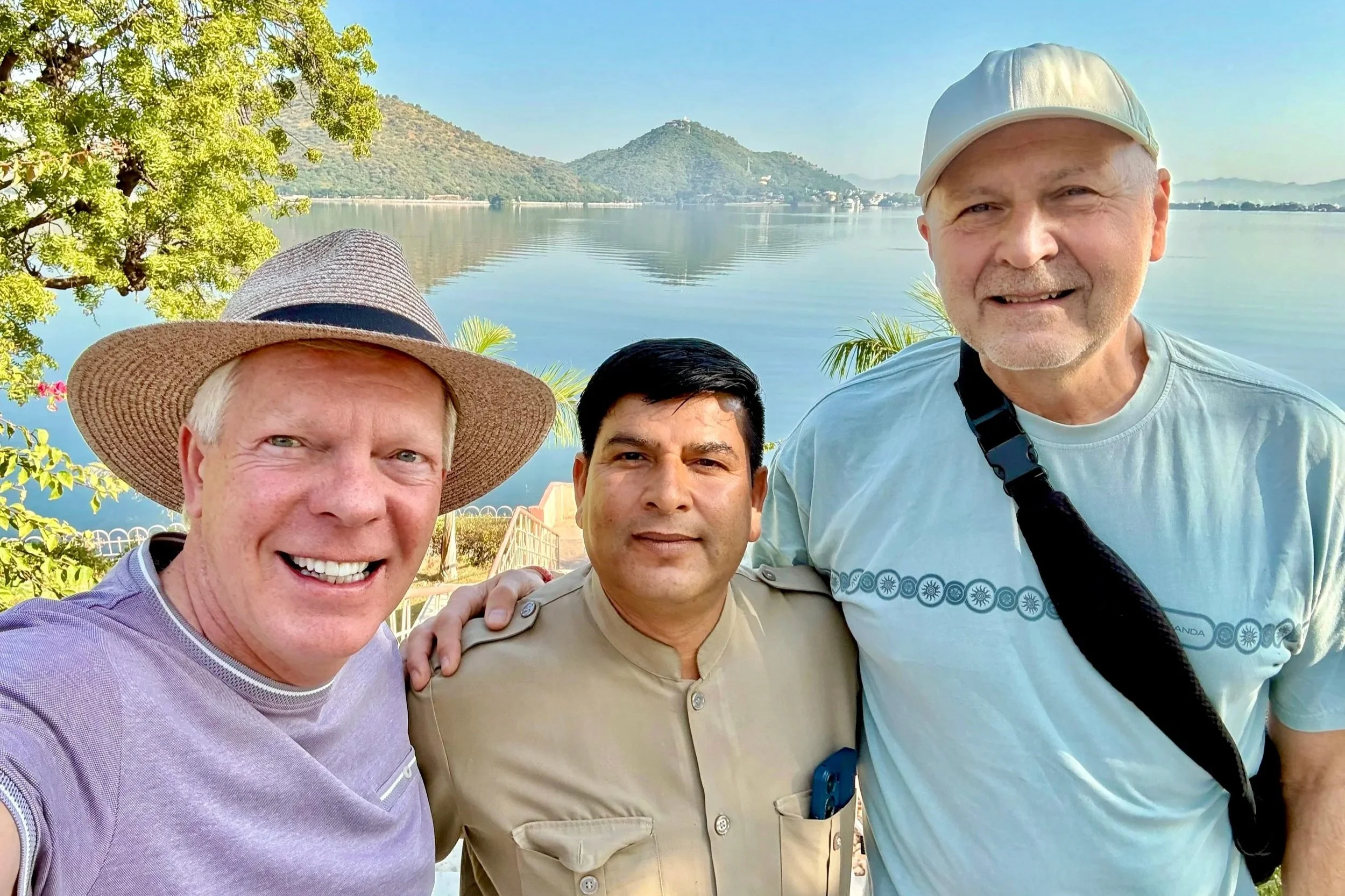 A group of four smiling men taking a selfie by a large body of water with mountains in the background. One man is wearing a wide-brimmed hat, another a light-colored cap, and another a uniform.