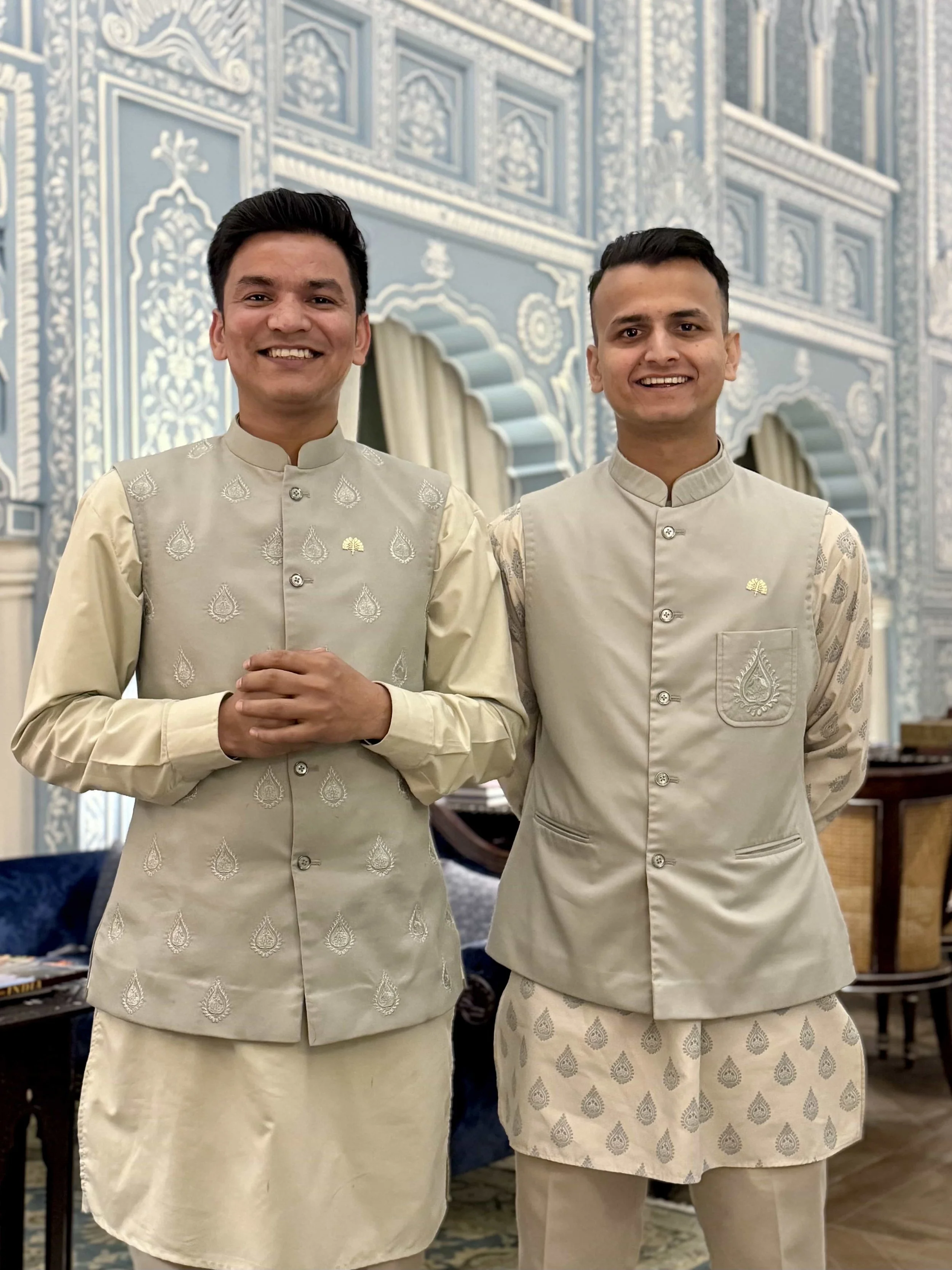 Two men dressed in traditional cream-colored Indian attire, standing inside a decorated building with intricate white and blue wall designs.