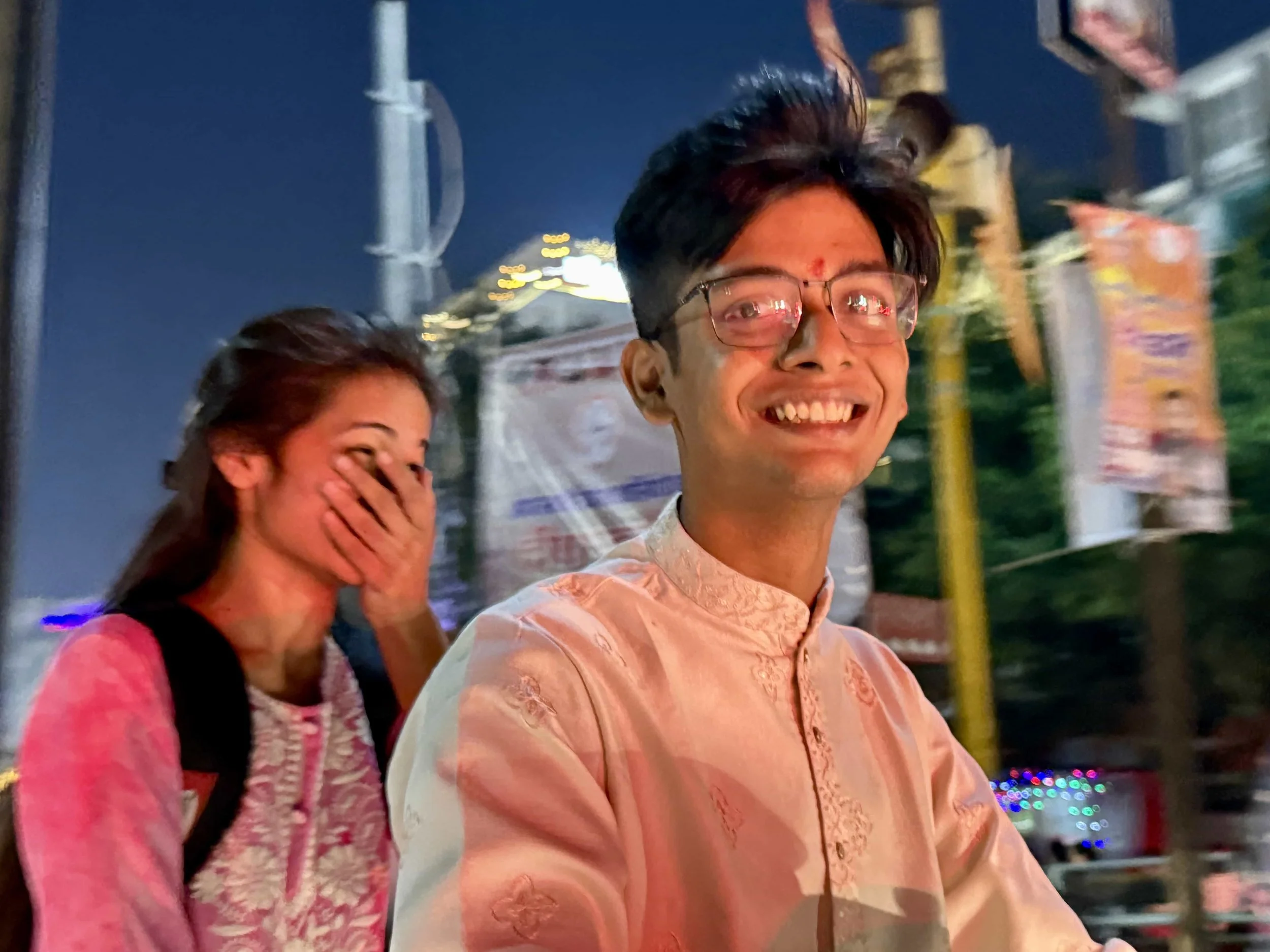 A young man wearing glasses and traditional clothing smiling at a nighttime festival, with a woman partially covered in pink and white traditional attire laughing nearby, and blurred colorful lights in the background.
